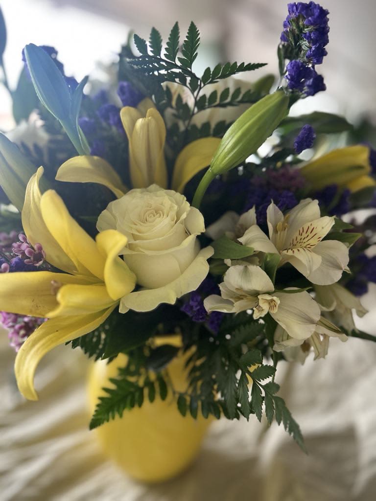 Sophia - Experience the vibrant beauty of our exquisite floral arrangement featuring a stunning combination of yellow lilies, white roses, and white alstroemeria, complemented by the rich hues of purple statice and purple wax flowers. This elegant bouquet is artfully designed in a charming yellow pitcher, making it a perfect centerpiece for any occasion. The harmonious blend of colors and textures not only brightens up your space but also conveys a message of joy and celebration, making it an ideal gift for loved ones or a delightful treat for yourself.
