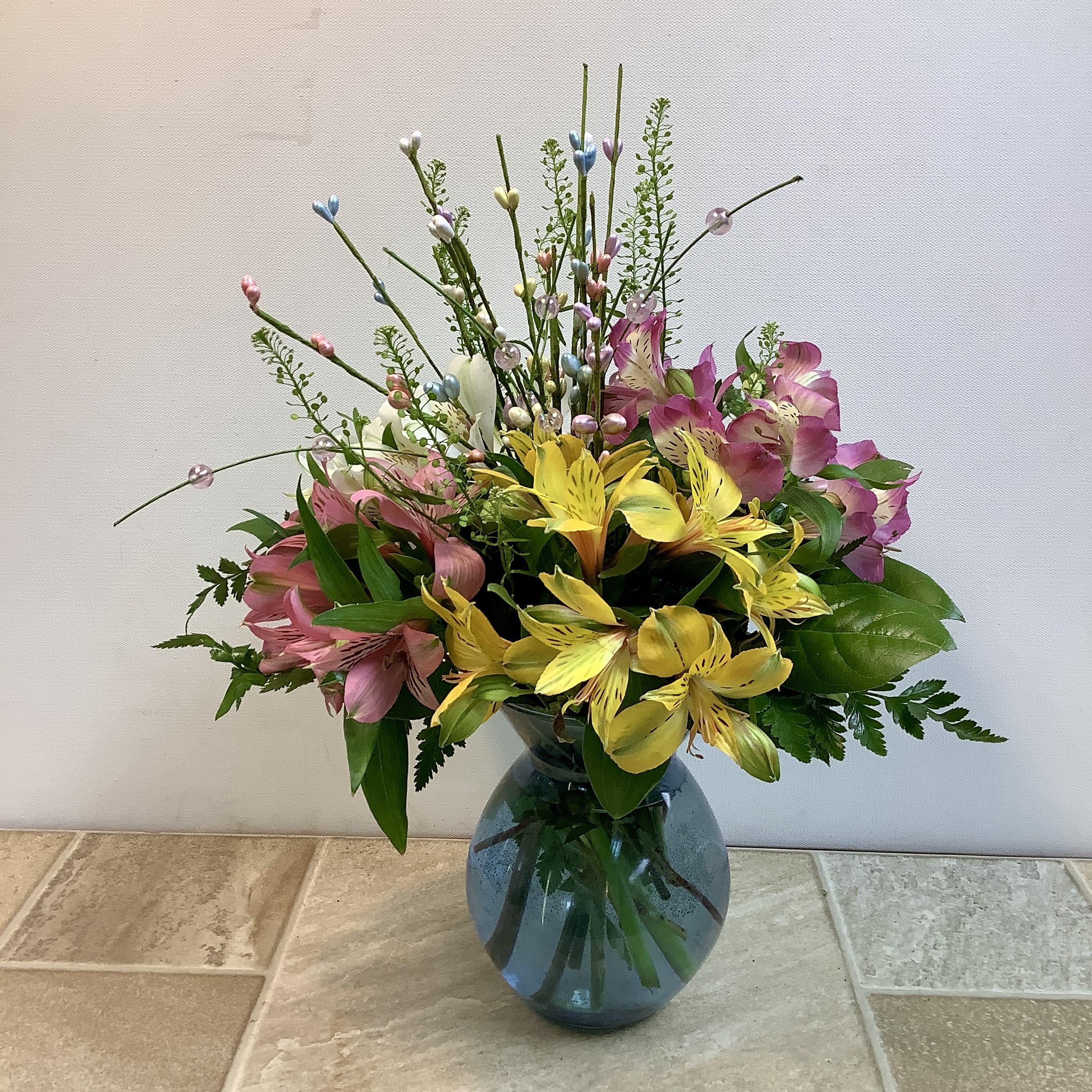 Easter Morning  - This Easter bouquet is a joyful celebration of spring’s soft colors and playful textures. Bright yellow, pink, lavender and white alstroemeria bloom generously at the heart of the arrangement, their delicate speckled petals adding warmth and charm. Airy greenery and looping grass tips dance upward, adorned with pastel bead accents in soft blues, pinks, and lavenders—reminiscent of Easter eggs glistening in the morning light. Arranged in a rounded blue glass vase, the design feels fresh, light, and full of seasonal cheer. It measures 11” wide x 17” tall.