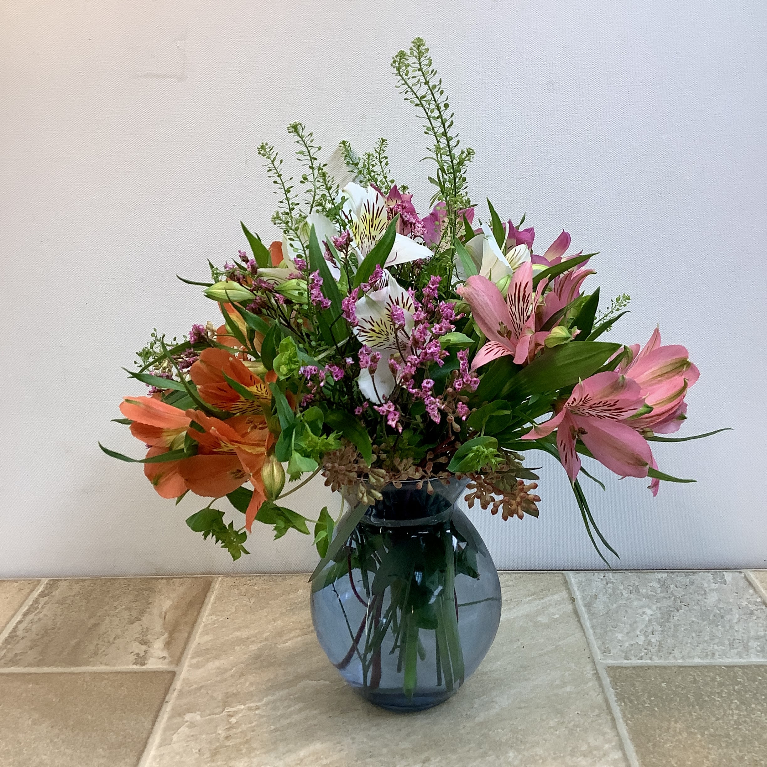 Pastel Jubilee  - This bouquet is a bright and lively mix bursting with color and movement. Pastel shades of alstroemeria create a warm, cheerful palette. Sprigs of delicate pink limon weave throughout, adding texture and a touch of whimsy, while airy green filler and fresh foliage give the arrangement height and a garden-gathered feel. Designed in a rounded blue glass vase, the stems are visible through the water, adding a modern, clean finish. The overall look is fresh, joyful, and naturally styled — like a handful of blooms gathered on a sunny afternoon and arranged with effortless charm. It measures 12” wide x 16” tall.