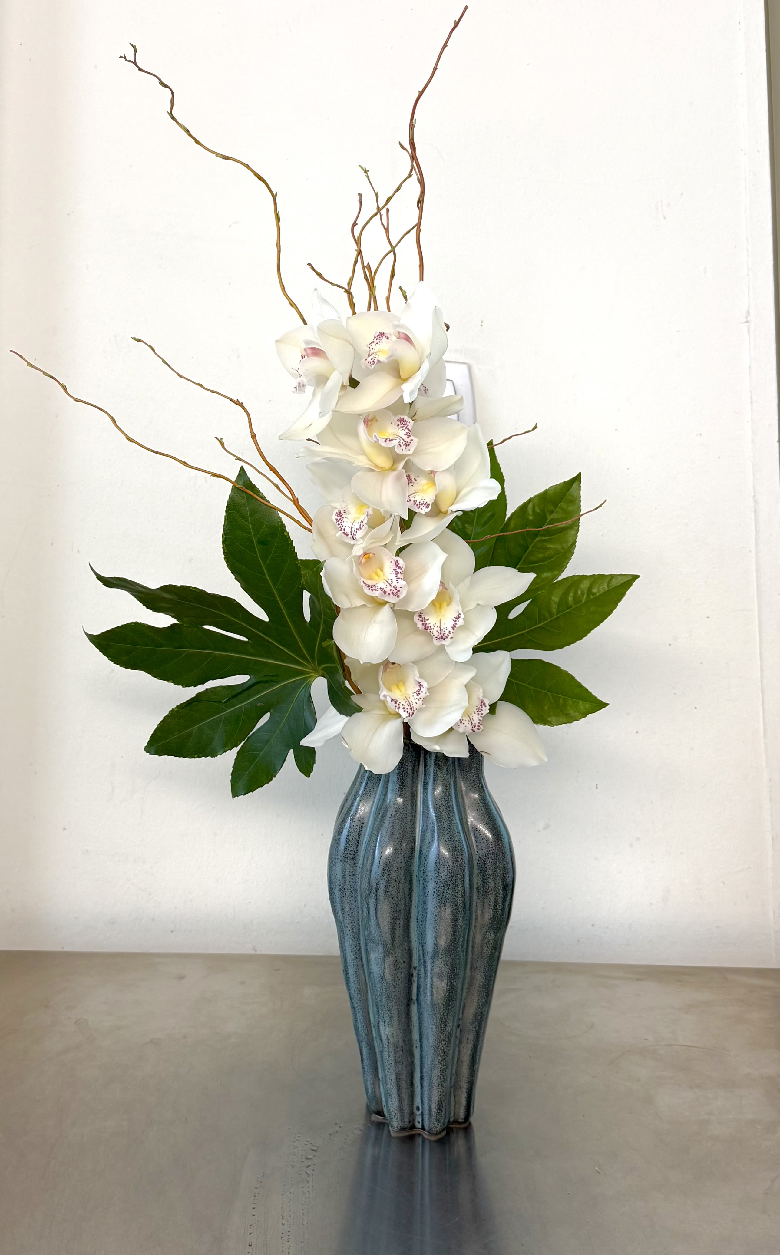 Orchid Cascade Arrangement - A striking vertical arrangement of elegant white cymbidium orchid designed to create a sense of movement and sophistication. Layers of soft orchid blooms rise gracefully among natural curly branches, complemented by bold tropical foliage for contrast and texture. Arranged in a sculptural ceramic vase with a deep blue glaze, this piece blends modern design with organic beauty.  Perfect for entryways, offices, or special gifts, the Orchid Cascade brings a refined, contemporary touch while highlighting the timeless elegance of orchids. 