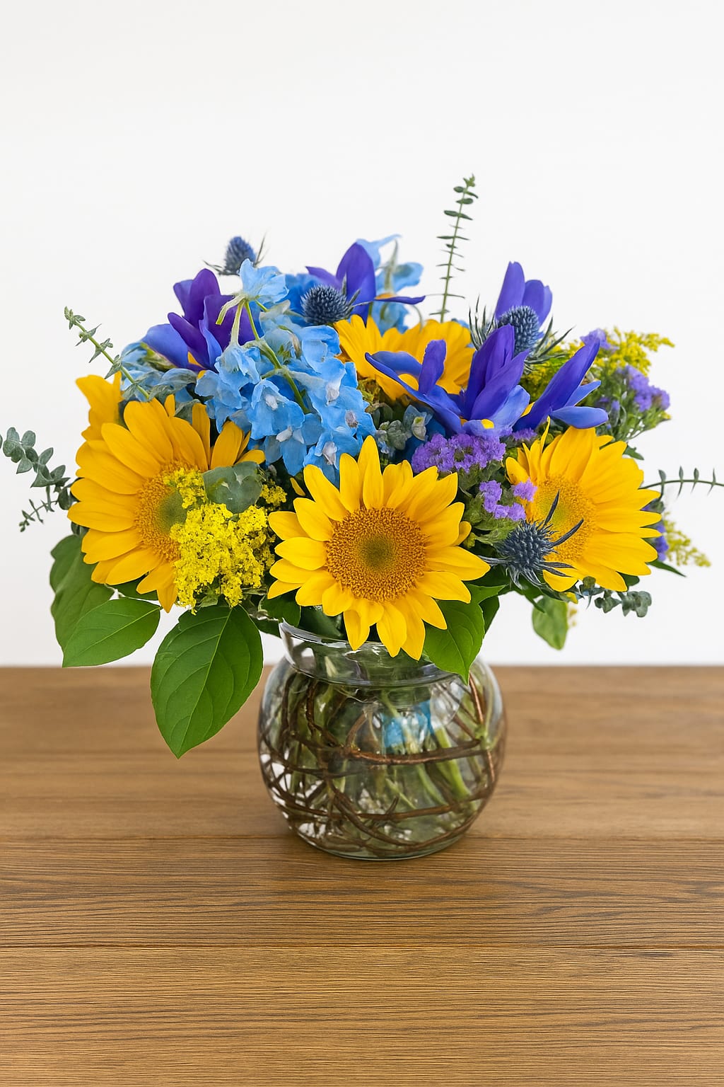 Sun and Sky - Arranged in a bubble bowl, this serene sun and sky design features fresh cut sunflowers nestled amidst a lush collection of blue thistle, iris, and belladonna and is accented with yellow solidago and eucalyptus.