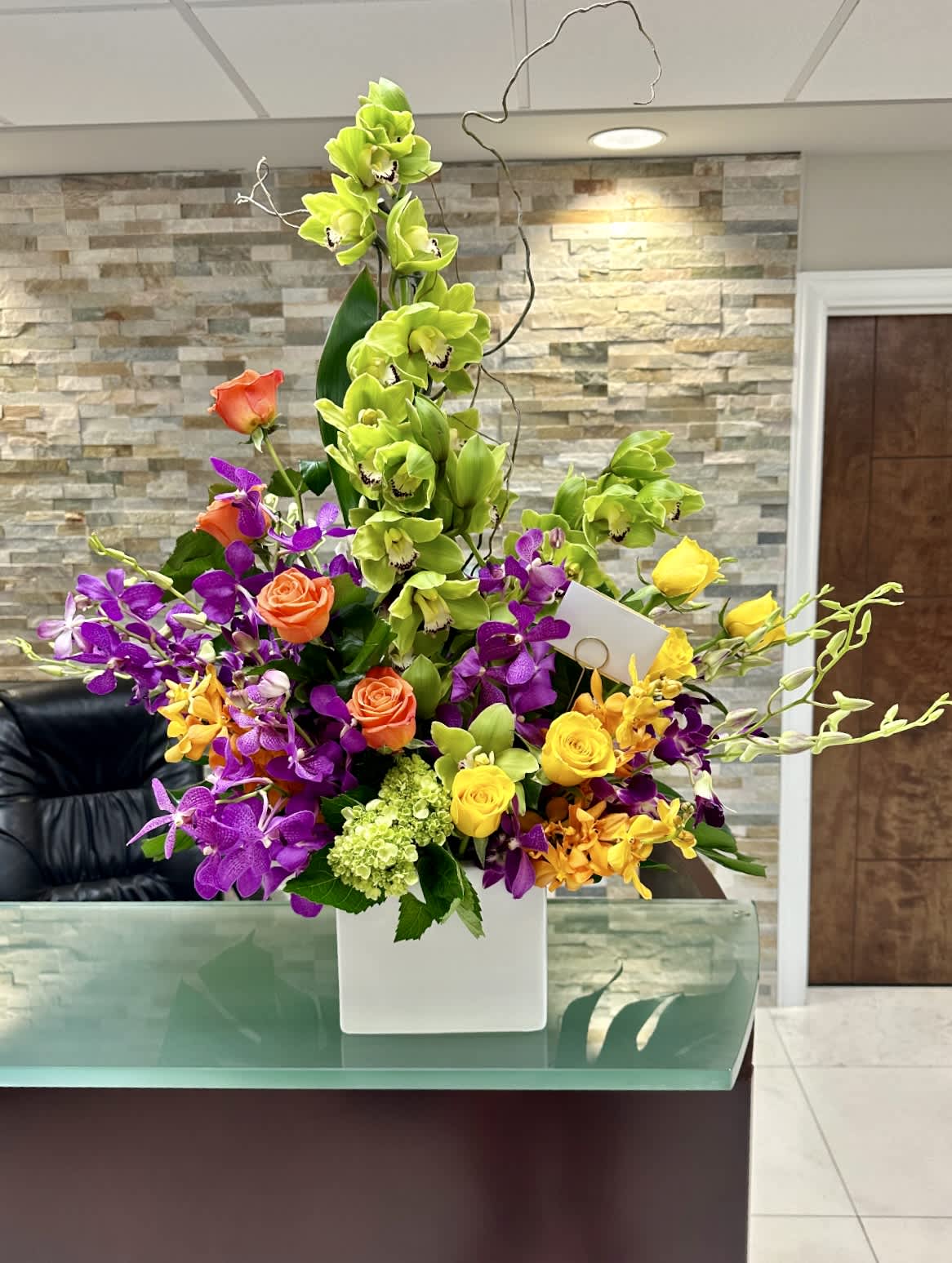 Spontaneous ™ - A statement piece overflowing with vibrant color—orange and yellow roses, green cymbidium orchids, purple orchids, and golden accents. Designed in a tall white vase, it’s perfect for lobbies, events, or as a dramatic centerpiece.