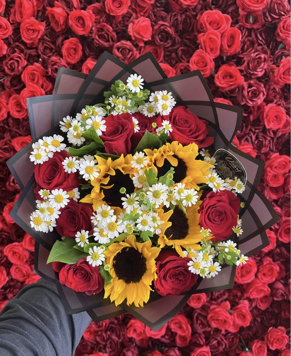 Handheld bouquet of red roses, yellow sunflowers, and white daisies wrapped in black paper over a bed of red roses.