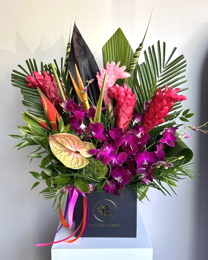Tall tropical arrangement with pink ginger, purple orchids, and anthuriums in a black box with ribbons