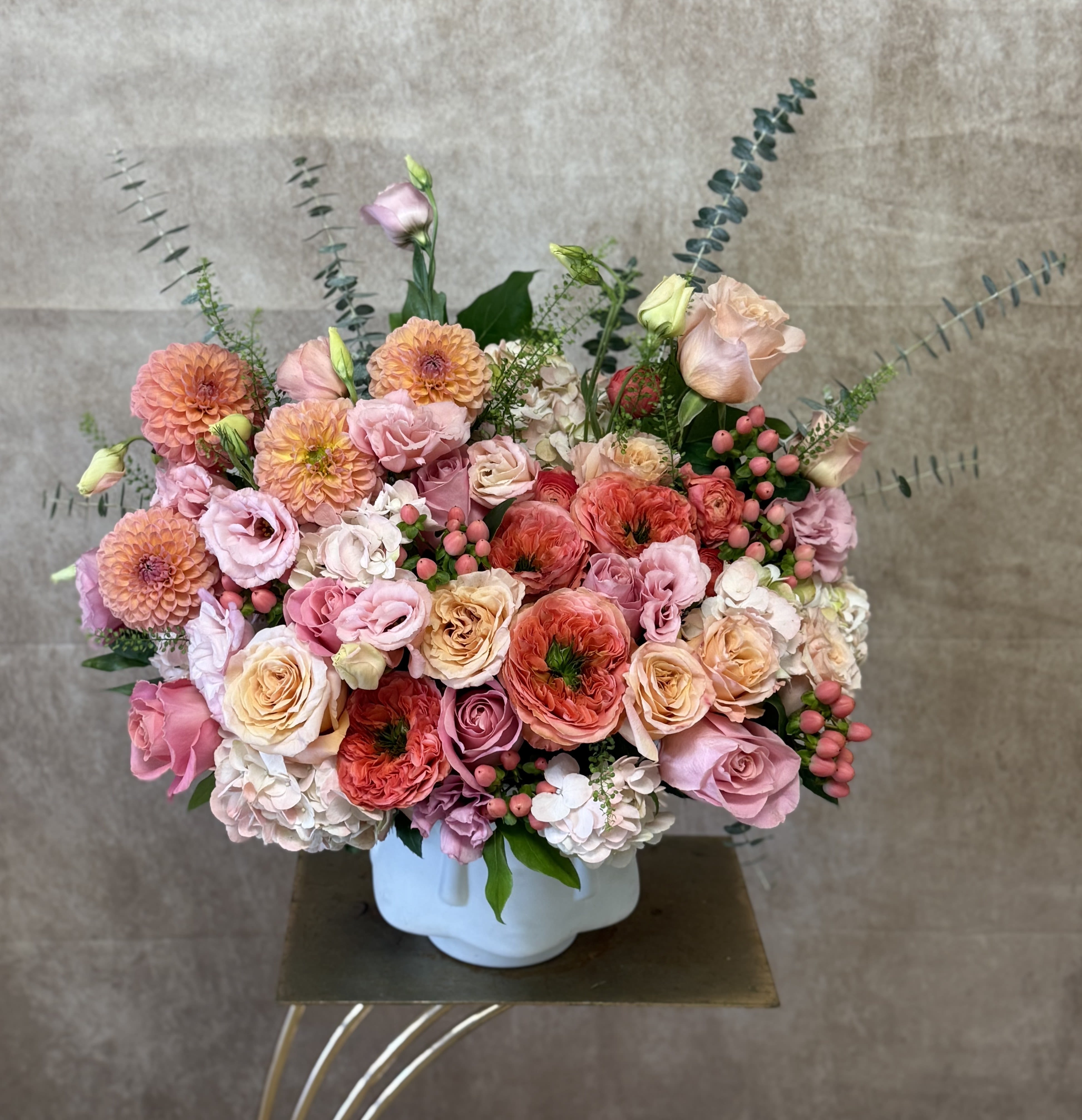 Gardens Paradise - Santa Clarita Valley Florist  - At the heart of this arrangement, dahlias reign with theatrical presence. Their intricate, geometric petals unfold in perfect concentric patterns, offering a stunning depth and dimension. From the bold, dinner-plate sized blooms to the charming, pom-pom varieties, they provide a captivating structural centerpiece in a painter’s palette of deep crimsons, blushing peaches, and vibrant magentas. Nestled amongst them are the impossibly romantic garden roses, the undisputed queens of fragrance and form. Their petals, softer and more numerous than classic roses, unfurl in a decadent, whisper-soft spiral. Each bloom is a masterpiece of subtle color gradation, with creamy ivory centers melting into blushing pink edges or apricot hues deepening to a copper kiss.