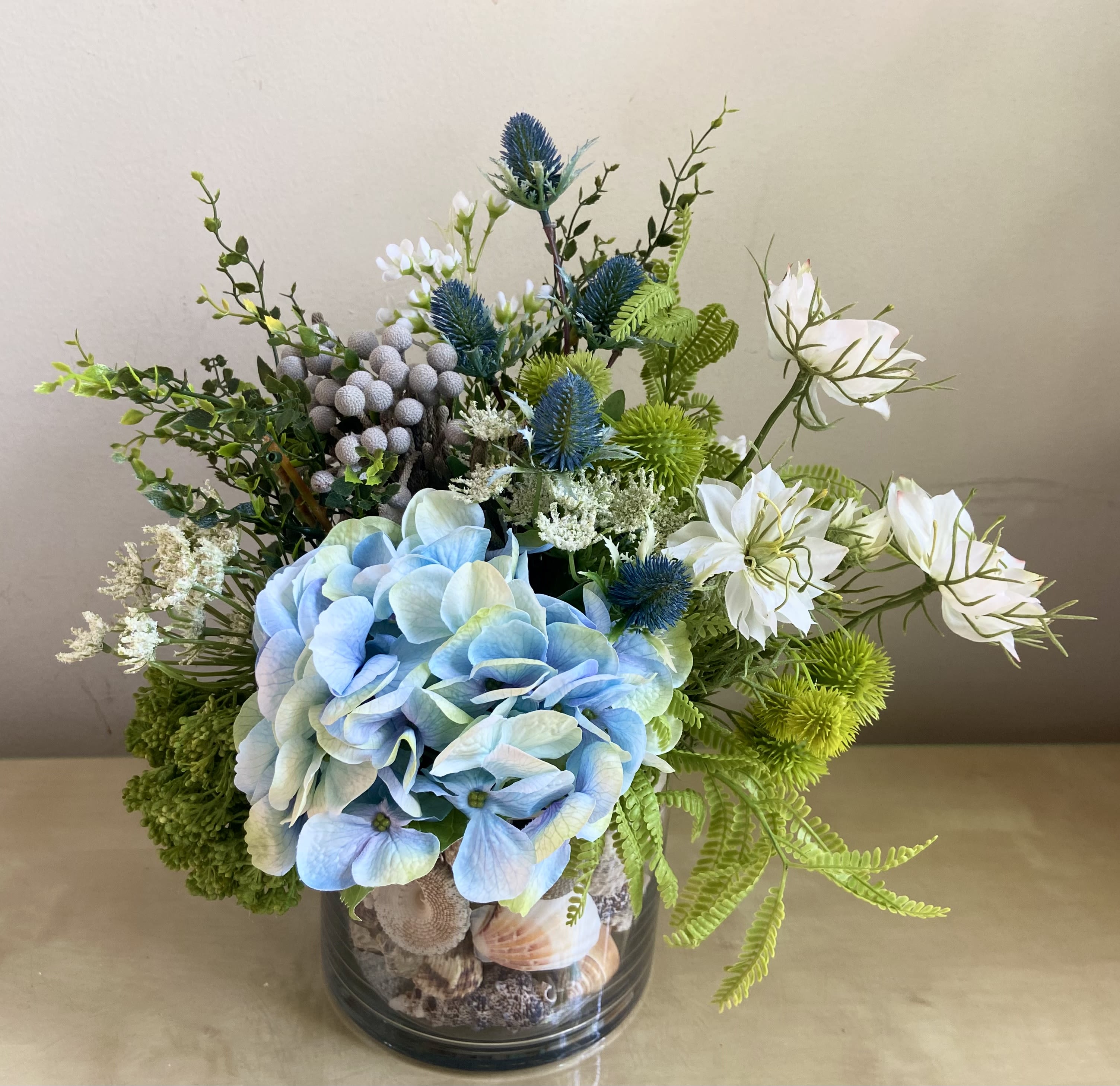 Cool Beach Vibe - Silk Floral Design - Warmer weather brings in the Cool Beach Vibe.  Lovely Silk floral arrangement in a bed of sea shells with blue hydrangea, nigella, thistle, quen anne’s lace, ferns and beach accents.