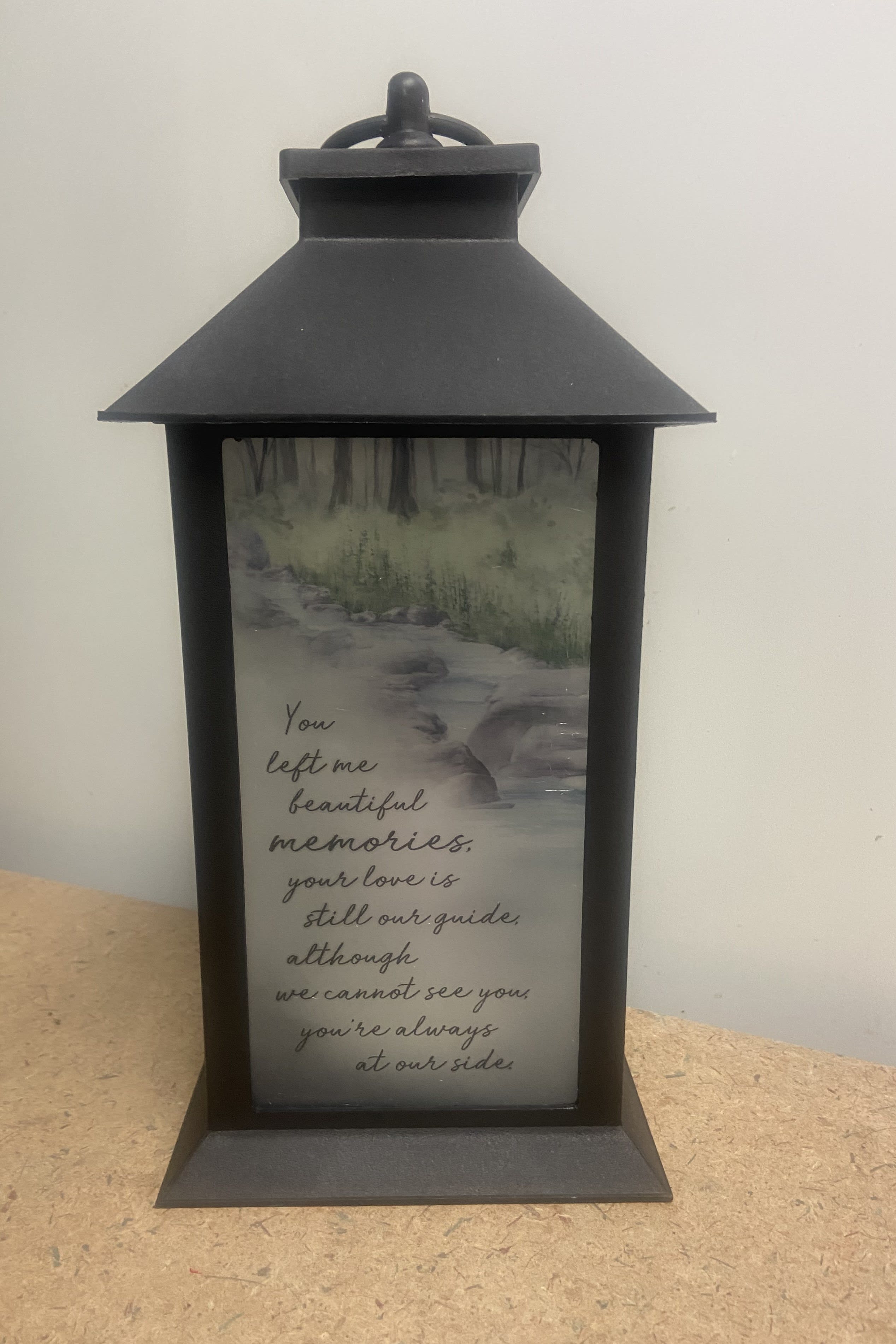 "Beautiful Memories" Lantern - This beautiful black high quality polyresin finish lantern surrounds an interior of a realistic LED flickering flame. The lantern has a "frost" look with a lovely mountain wooded stream pictured on the front panel  as well as the touching sentiment  "YOU LEFT ME BEAUTIFUL MEMORIES, YOUR LOVE IS STILL OUR GUIDE, ALTHOUGH WE CANNOT SEE YOU, YOU'RE ALWAYS AT OUR SIDE". Lantern is 13"H x 5 1/2"W x 5 1/2" D. A built in automatic timer and 3 AAA batteries are included. This item is suitable for sympathy gifting either at the vistation or the home  This item is weather friendly.