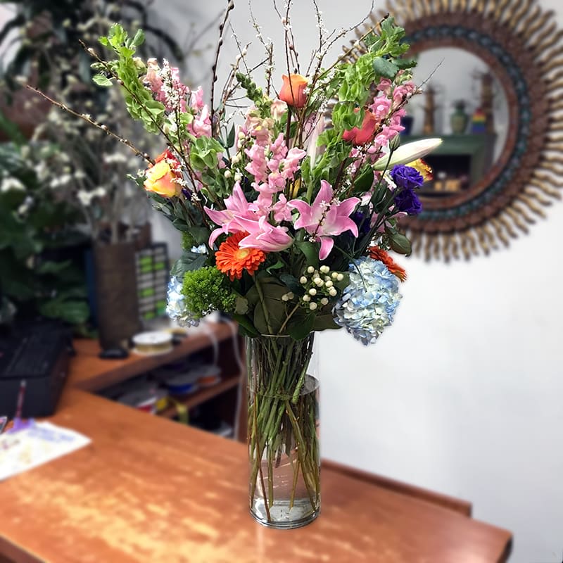 Sunrise in Brooklyn - Welcome to Brooklyn! An exclusive James Weir design to start your week. Two-tone yellow roses, orange gerbera daisies and roses, blue hydrangeas, purple lisianthus, peach snapdragons, green bells of Ireland, white hypericum, and willow. Cylinder glass vase.