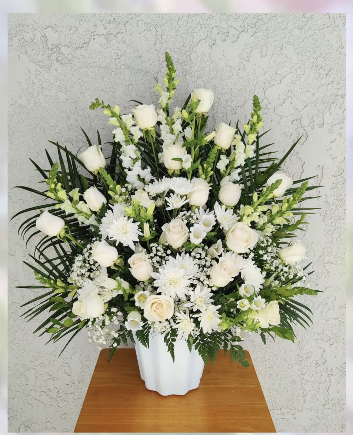 " Serenity " Sympathy Arrangement  - Express your deepest sympathies with this beautiful arrangement that radiates peace. It’s  created with harmonious flowers in white and green tones.  It includes: premium roses, snapdragons, cushion pompoms, fancy carnations, campanula bells, white filler flowers and seasonal foliage in a white vase .  One side arrangement. Large size   