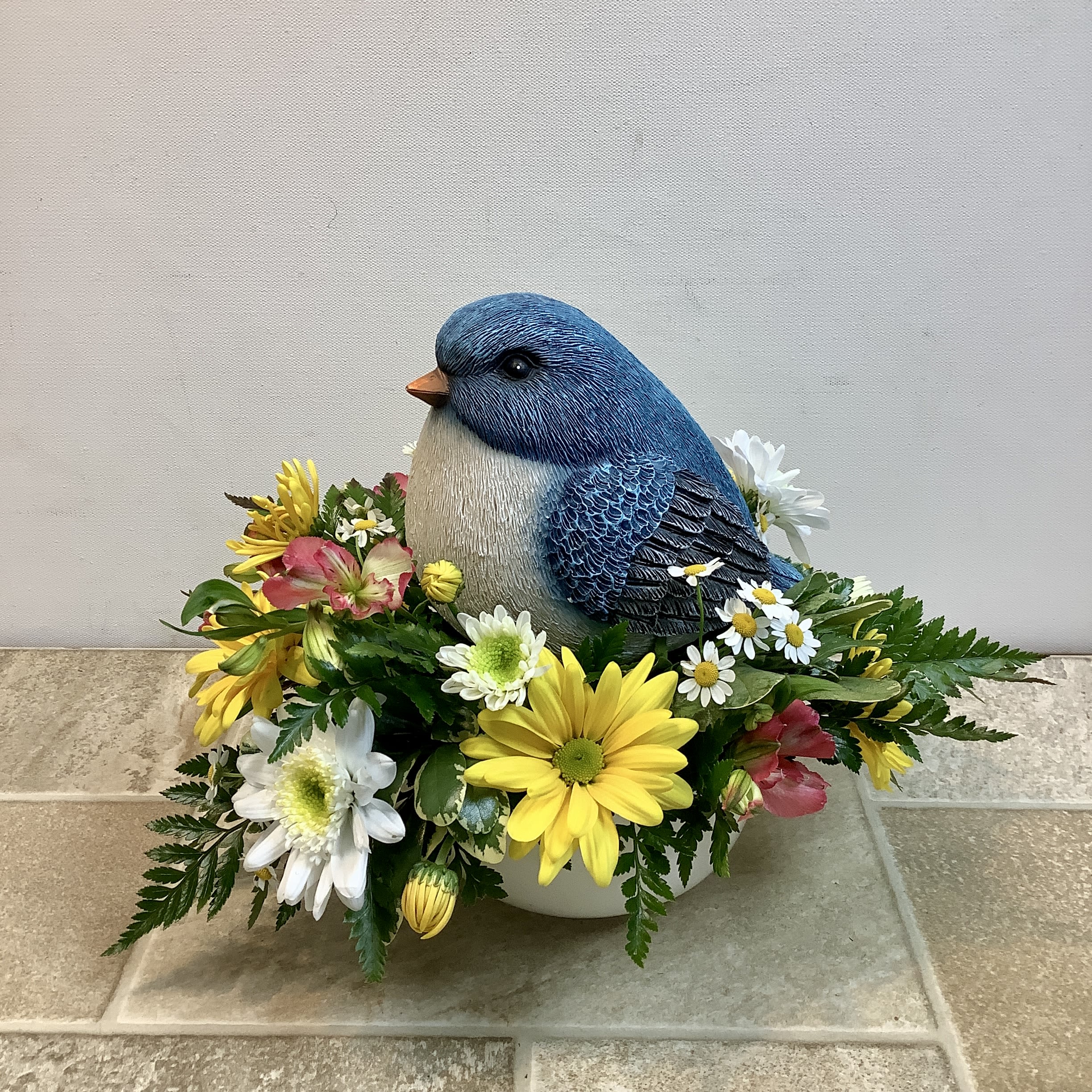 Bluebird of Happiness - This cheerful spring arrangement features a charming bluebird nestled in a bed of fresh blooms, creating the feeling of a tiny garden scene. Sunny yellow daisies, crisp white cushion mums, delicate chamomile-like daisies, and soft pink alstroemeria surround the keepsake bluebird, while lush greenery and fern accents add natural texture and fullness. Designed in a low container, this sweet arrangement brings the warmth and joy of a blooming garden and the promise of springtime indoors. It measures 11” wide x 10” tall.