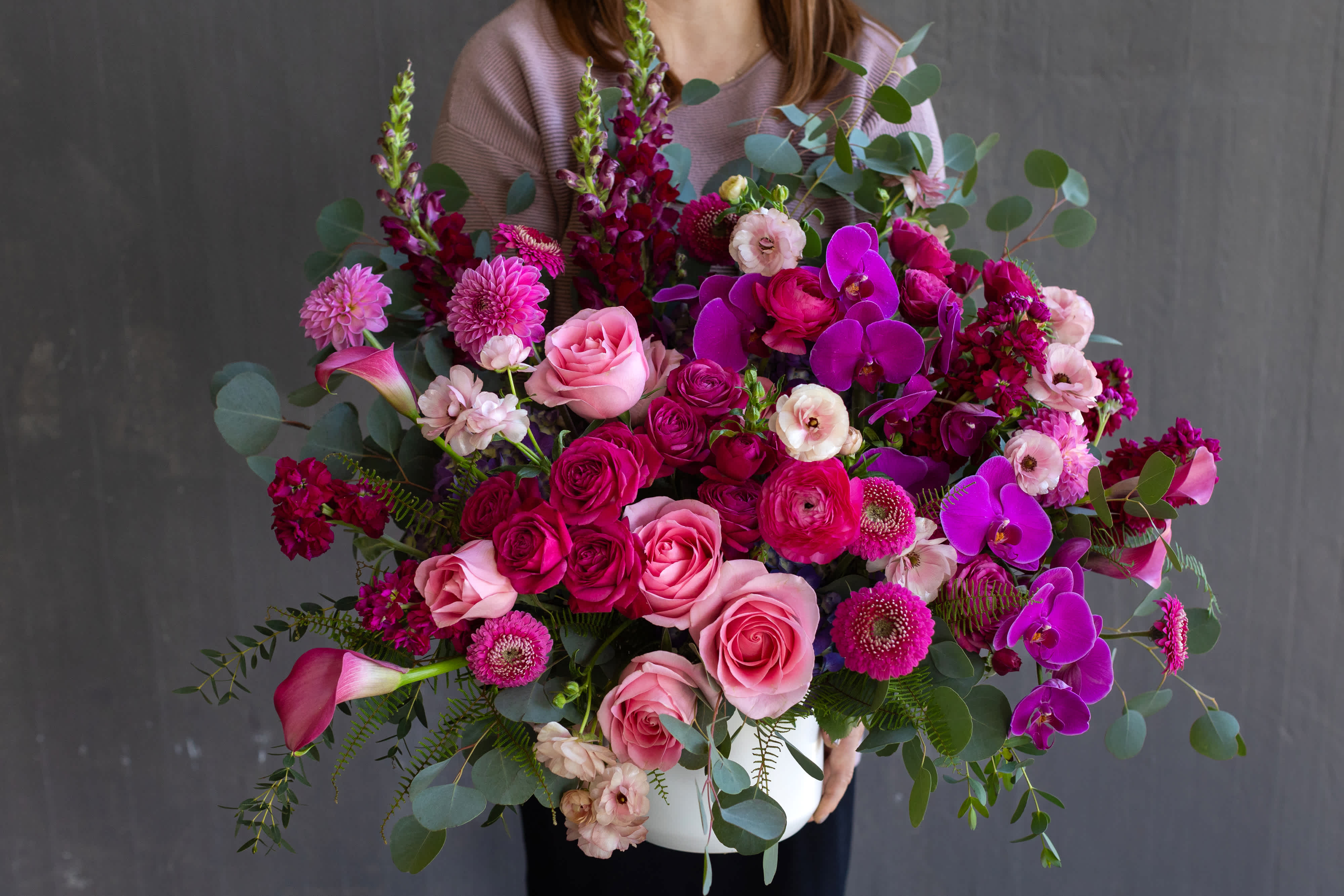 Fuchsia Delight- The Signature Collection - Dramatic and elegant. These showstopping fuchsia purple phalaenopsis orchids steal the show, delicately cascading atop the most vibrant pink roses! Designed in shades of purple and pink, this vase is abundant with blooms in a bursting and fun style. Our Fuchsia Delight flower arrangement is perfect for people who love luxury and a little theatrical drama.  This arrangement is offered as part of our Signature Collection. Due to the nature and scale of this design, we cannot offer same day delivery on any of our Signature Collection designs. We want to ensure the finest designs, and take care to ensure each arrangement is crafted with the highest quality flowers. Although we strive to create each arrangement as close to our photos as possible, there is always the chance that some flowers may be seasonally unavailable. In this case, we would substitute with flowers of a similar look and quality, without sacrificing the value of the design. Each arrangement will adhere to the color pallet and look of the arrangement ordered.     SIZE: This flower arrangement is approximately 20” height and 20” width.  FAQ Q: What time will my flowers arrive? A: Excellent question! For full details around our delivery process and timings, please see our Terms and Conditions. Q: May I request to change the design a bit? A: Yes, please make a note in the SPECIAL INSTRUCTION box when you place your online order. We will do our best to follow them.