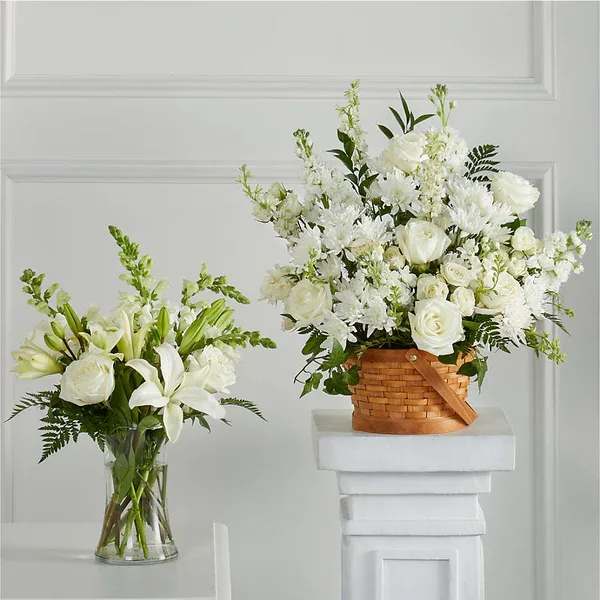 Two white floral arrangements with roses and lilies, one in a glass vase and one in a woven basket on a pedestal