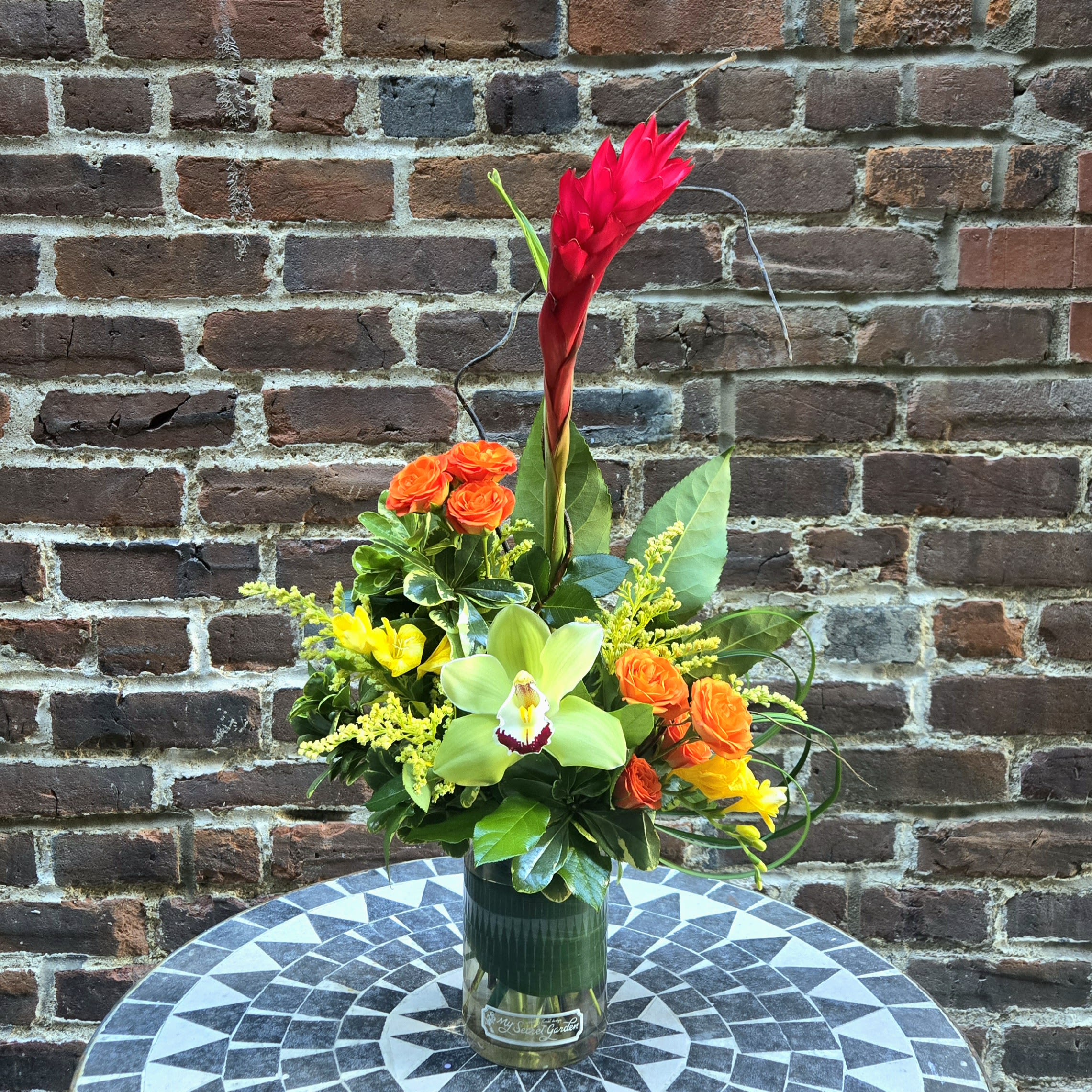 Tropical Sophistication - Tropical Sophistication is a stylish and unique floral design. Presented in colors of red, orange, yellow, and green, this stunning design includes red ginger, freesia, orange spray roses, and solidago accented with curly willow and tropical foliage's. A wonderful design perfect for any home or office space. 