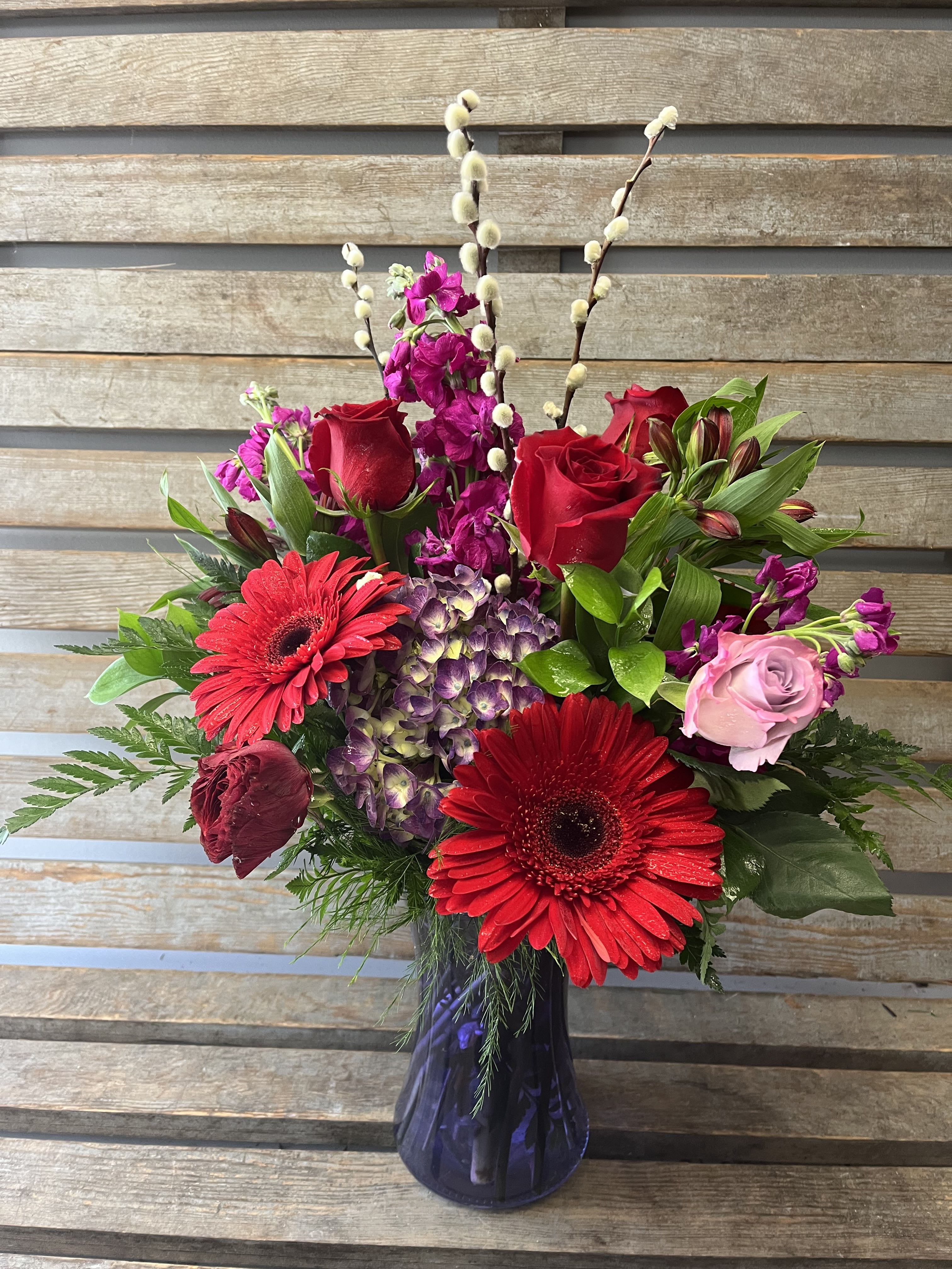 Jewel tone bold color arrangement  - Rich reds, purples , fuschia in colored glass vase. Designer’s choice of flowers. Different each week. Pussy willow when available. 