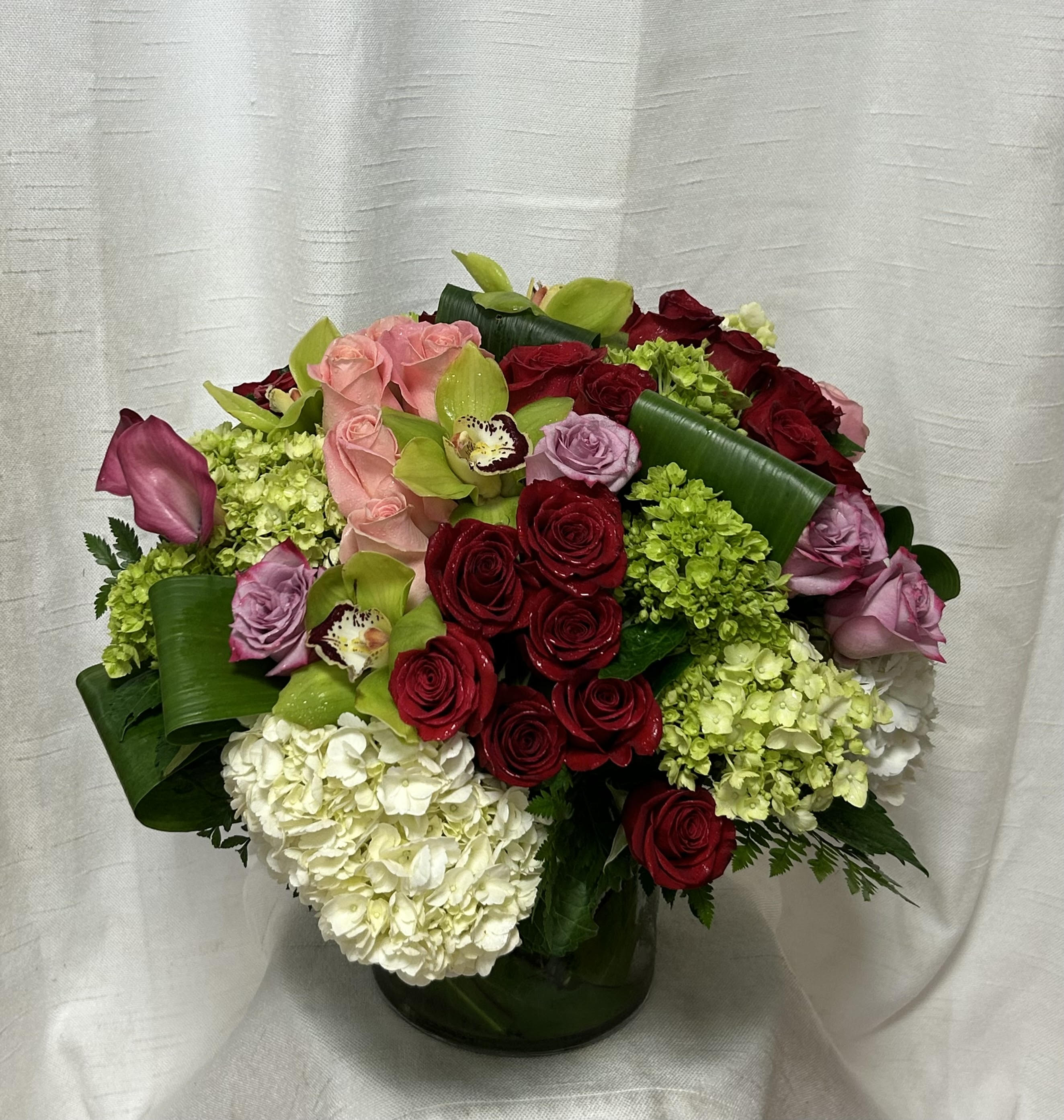 Romanced (DB) - Elevate any space with our exquisite and upscale floral arrangement. Impeccably designed by our expert florists, this stunning arrangement features a striking combination of green and white hydrangeas, red roses, pink tulips, and pink orchids, beautifully arranged in a large 7x7" leaf-lined glass cylinder vase.
