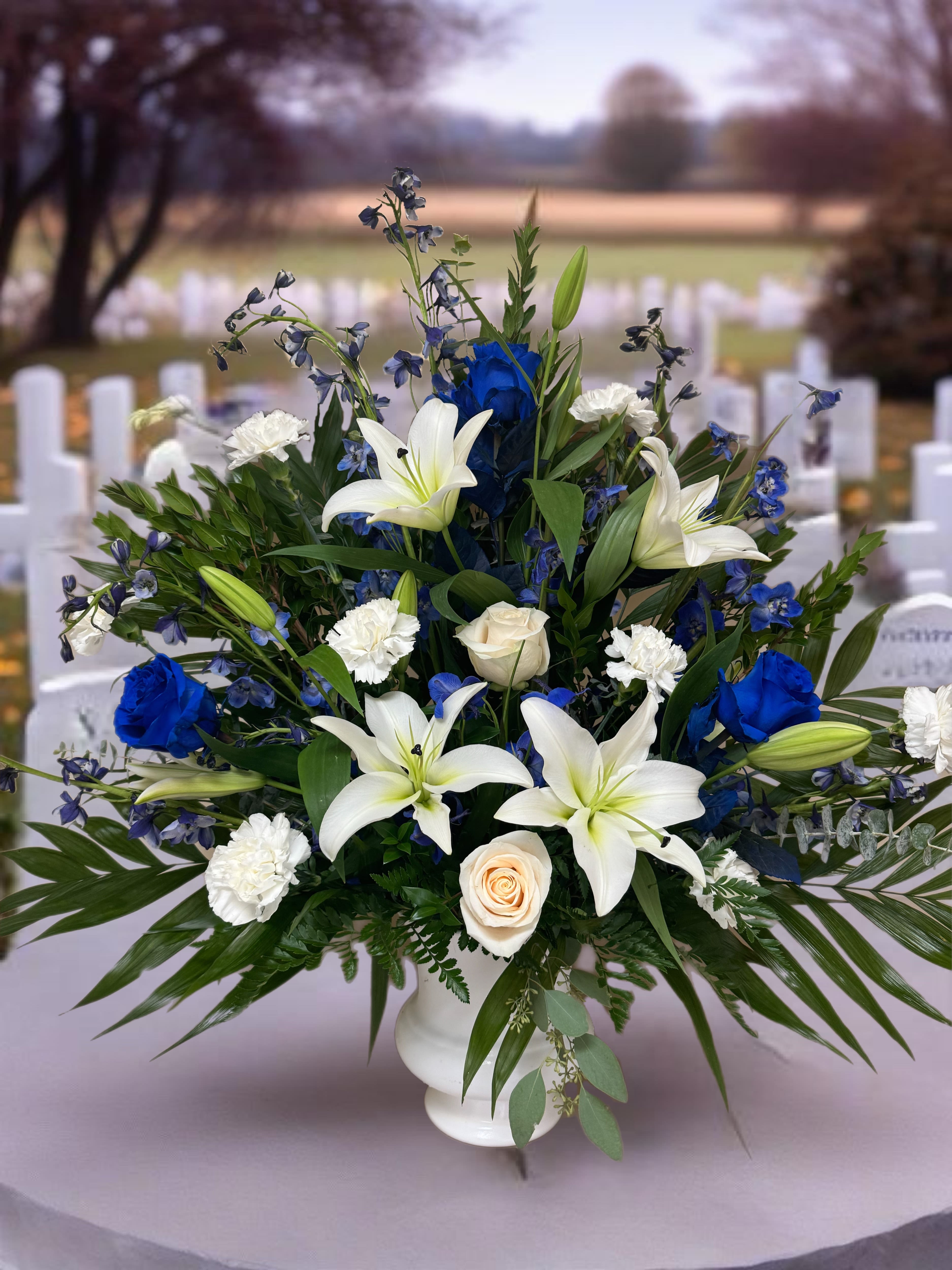 Celestial Tide - A striking blend of blue and white Roses represents unwavering devotion and peaceful farewells, while pure white lilies symbolize the light of cherished memories. Soft carnations add depth and texture, while delicate blue delphinium reaches skyward, evoking a sense of ascension and eternal peace. This arrangement serves as a heartfelt tribute, mirroring the beauty of the horizon where the earth and sky unite. (This pairs well with the "Blue Horizon" casket cover and either the "Stunning Sapphire" easel or "Serenity" easel)