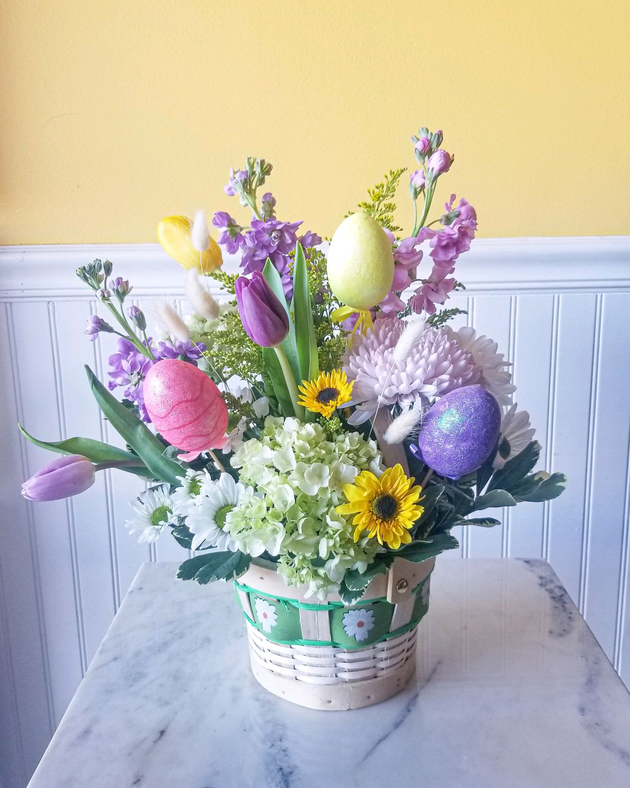 Easter Basket - A festive woodchip basket filled with Easter spring blooms and accents. Basket color may be green, pink, blue or purple (based on availability).  Approx dimensions 14 in W X 16 in H *Note arrangement is 1 sided*