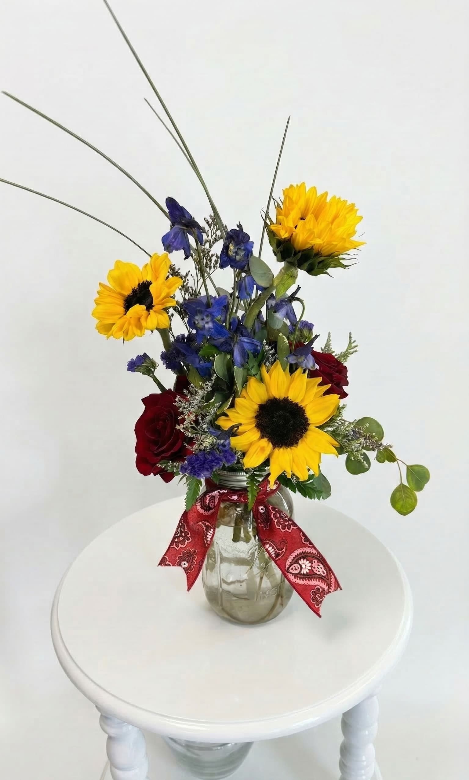 Hill Country Harmony - A Texas Classic for Any Celebration "Bring the soulful beauty of the Texas Hill Country right to their doorstep. Hill Country Harmony is a vibrant, textured arrangement that balances the bold, sunny energy of premium sunflowers with the deep, romantic velvet of red roses. Touches of midnight blue delphinium add a striking contrast, all gathered in a timeless mason jar and hand-finished with our signature red paisley bandana.  Whether you’re celebrating a milestone birthday, sending a heartfelt 'thank you,' or simply brightening a friend's day, this arrangement captures a spirit of warmth and authentic charm that fits every occasion perfectly."