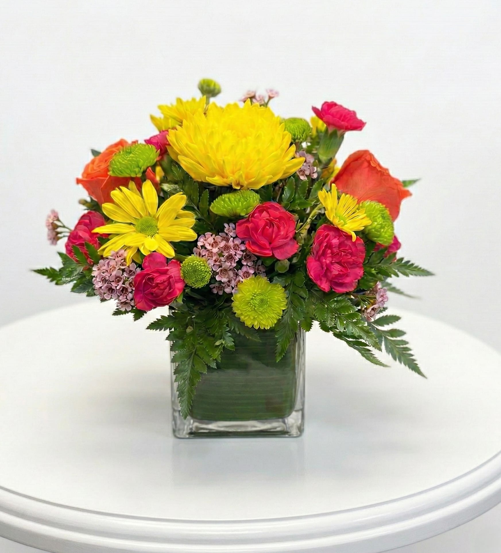 Touch of Sunshine Bouquet - Colorful blooms that are ready to brighten your recipient's day. This flower bouquet is bright, happy and ready to celebrate any occasion! Orange roses and yellow Asiatic Lilies are vibrant and fun surrounded by yellow daisies, hot pink mini carnations green button poms, filler flowers and an assortment of lush greens presented in a clear glass cubed vase lined with ti leaf green material for added beauty this unforgettable bouquet is that ultimate surprise that will make them feel the love on any day of the week.  