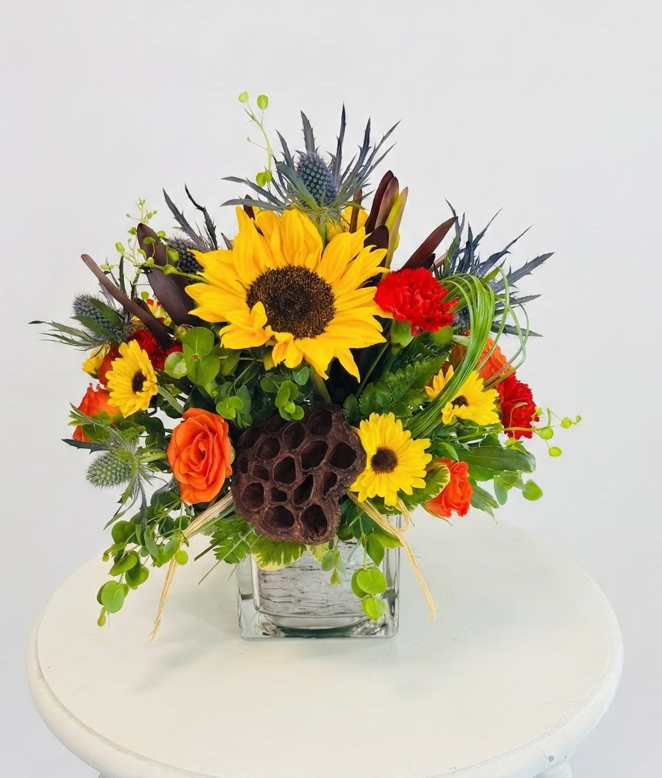 Artisan's Edge Bouquet - Where botanical architecture meets vibrant color. This design plays with height and texture, pairing the soft velvet of sunflowers and roses against the rugged, sculptural beauty of a dried lotus pod and spiky sea holly. Designed in a compact cube, "Sun-Drenched Textures" is a sophisticated choice for a desk or side table, offering a 360-degree view of nature’s most interesting shapes.