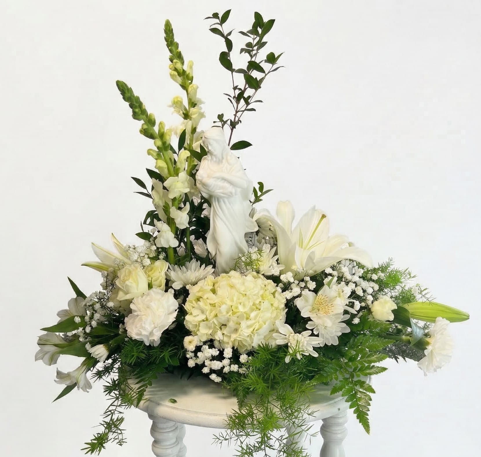 Heavenly Serenity Bouquet - A breathtaking all-white masterpiece that combines texture and height to create a focal point of quiet reflection. Soft white Hydrangeas and delicate Alstroemeria provide a cloud-like base for the elegant Madonna centerpiece, while architectural Snapdragons draw the eye upward. Perfectly balanced with fresh greenery and airy accents, this arrangement is a sophisticated choice for services, anniversaries, or religious milestones.  (Includes Keepsake Figurine)