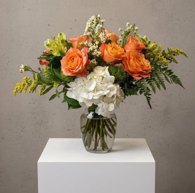 Golden Glow - Radiant and uplifting, Golden Glow is a bright arrangement that brings warmth and elegance together. Featuring vibrant orange roses as the focal point, this design is softened by a lush cluster of white hydrangea and accented with alstroemeria, delicate white stock, and touches of golden solidago. Fresh greenery and textured foliage add depth and movement, all beautifully arranged in a classic clear glass vase.