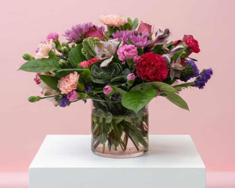 Wildflower Bliss - Playful, colorful, and full of texture, Wildflower Bliss is a charming garden-style arrangement that feels fresh and naturally abundant. This design features a lively mix of pink and lavender mums, soft peach carnations, vibrant red carnation, delicate alstroemeria, and touches of purple statice. At the center, a unique succulent accent adds a modern, earthy touch, while lush greenery and heart-shaped foliage bring movement and balance. Arranged in a clear glass cylinder vase, this bouquet captures the relaxed beauty of a blooming wildflower garden.