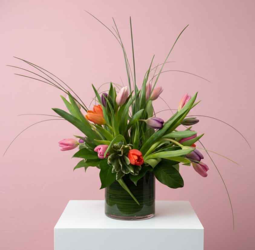 Spring Breeze - Fresh, modern, and full of movement, Spring Breeze captures the light and airy feeling of the season. This elegant arrangement features a vibrant mix of pink, purple, orange, and coral tulips surrounded by lush greenery and flowing ornamental grasses that add height and graceful texture. Designed in a sleek glass cylinder vase lined with green foliage, the look is clean, contemporary, and naturally beautiful.
