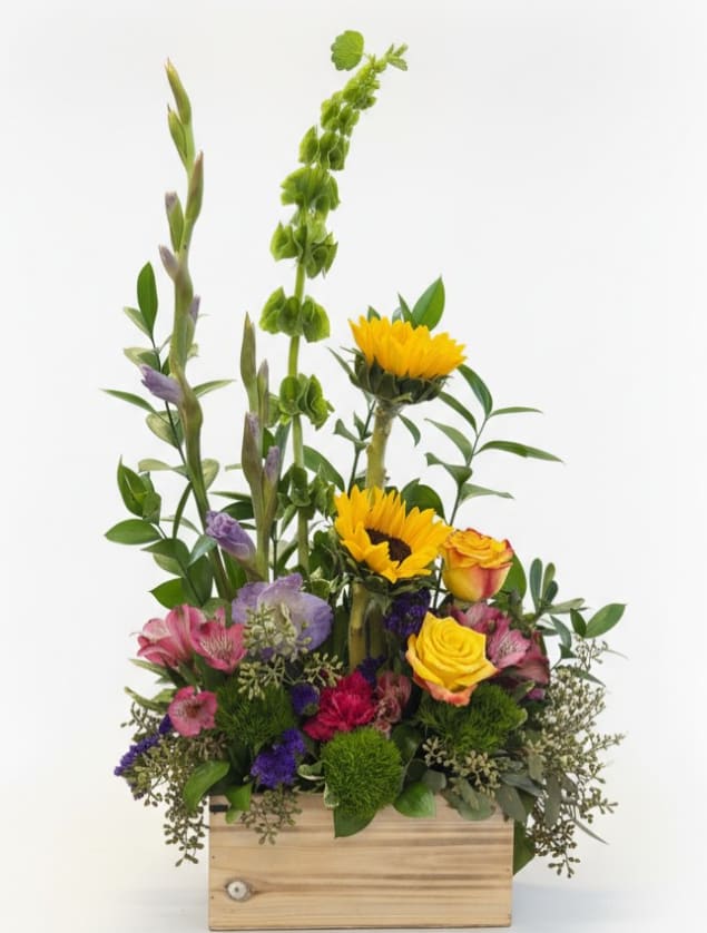 Sunny Meadow - Bright, cheerful, and full of natural charm, Sunny Meadow brings the beauty of a blooming countryside garden indoors. This unique design features radiant sunflowers paired with golden yellow roses, soft pink alstroemeria, and vibrant purple and green accent flowers for texture and color. Tall stems add graceful height while lush greenery surrounds the blooms, all beautifully arranged in a rustic wooden planter box that enhances its fresh-from-the-garden feel.