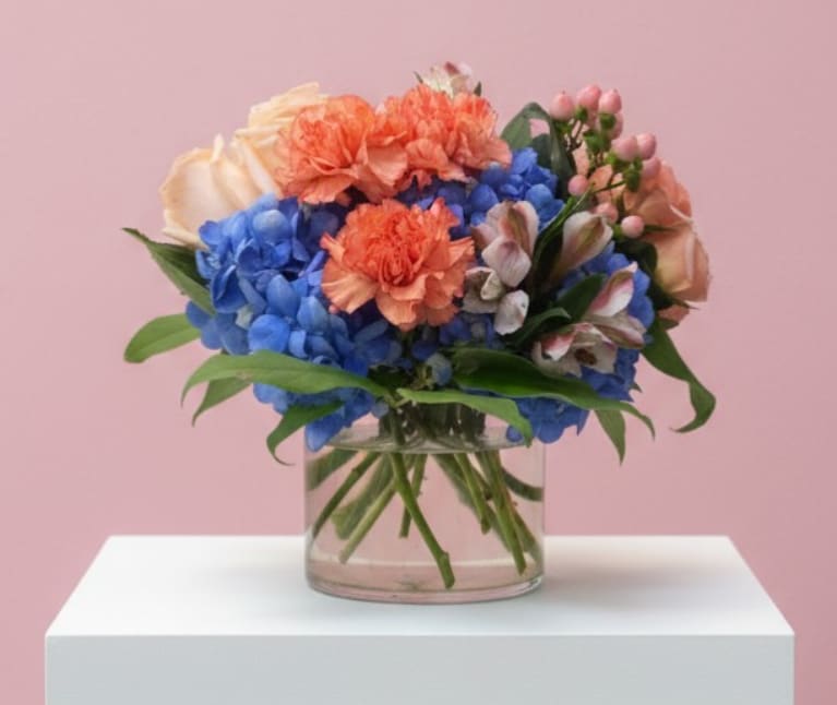 Coastal Charm - Soft, colorful, and effortlessly elegant, Coastal Charm blends vibrant blues with warm peach tones for a fresh and uplifting look. Lush blue hydrangea forms the base of this arrangement, beautifully complemented by peach roses, delicate coral carnations, soft alstroemeria, and accents of pink hypericum berries. Fresh greenery surrounds the blooms, all arranged in a modern clear glass cylinder vase for a clean and contemporary finish.
