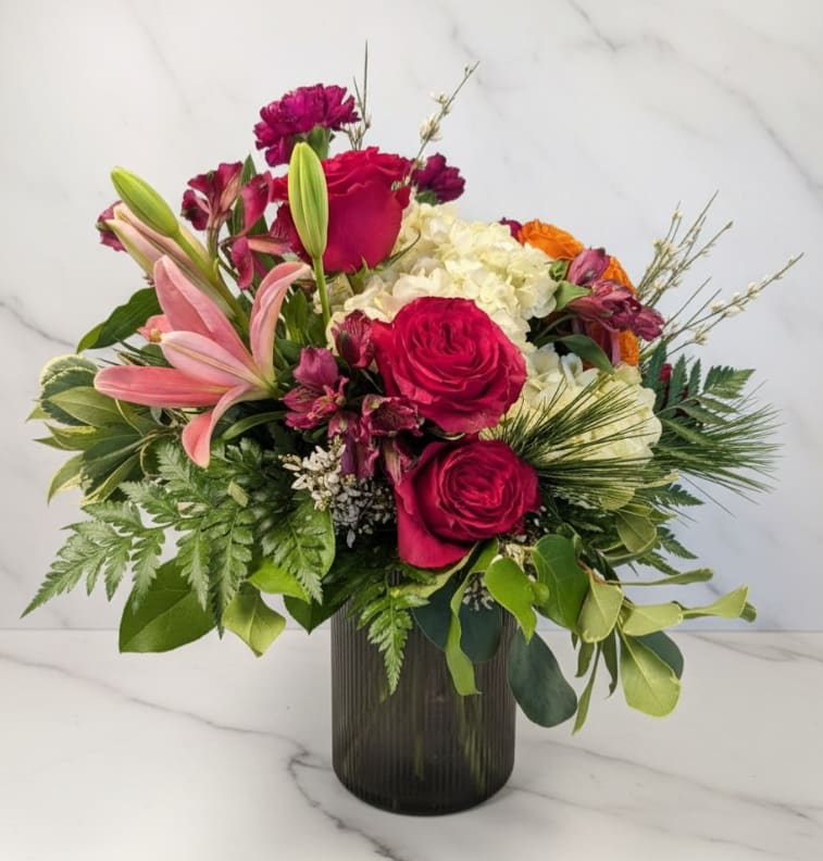 Ruby Garden - Bold, romantic, and full of lush texture, Ruby Garden is a rich floral arrangement designed to make a lasting impression. Vibrant red roses take center stage, beautifully complemented by a graceful pink lily, soft white hydrangea, and delicate alstroemeria. Seasonal greenery and textural accents add depth and movement, all arranged in a sleek modern black glass vase that enhances the dramatic color palette.