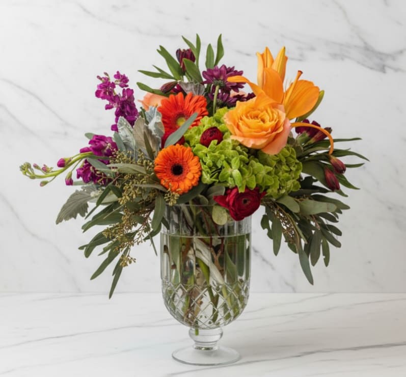 Autumn Radiance - Warm, vibrant, and full of rich seasonal color, Autumn Radiance is a stunning arrangement that captures the beauty of a glowing fall garden. A lush green hydrangea sits at the center, surrounded by vibrant orange roses, cheerful gerbera daisies, bold orange lilies, and deep red ranunculus. Accents of purple stock and textured greenery add depth and movement, creating a beautifully balanced design. Arranged in an elegant cut crystal-style pedestal vase, this bouquet feels both luxurious and full of natural charm.