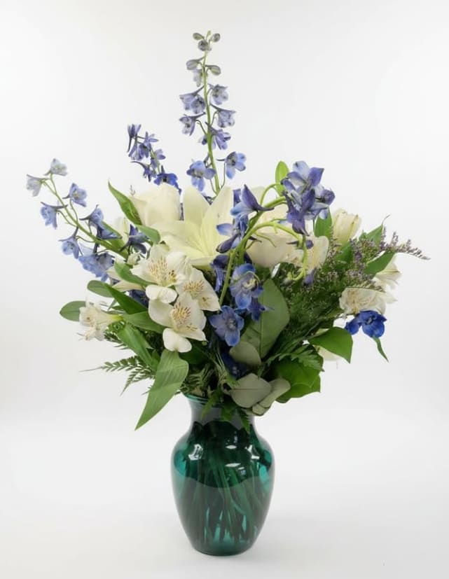 Blue Serenity - Graceful and calming, Blue Serenity is a beautifully balanced arrangement that blends soft whites with cool blue tones. Elegant white lilies bloom among delicate blue delphinium, creating height and natural movement. Accents of white alstroemeria and lush seasonal greenery add texture and softness, all arranged in a rich emerald green glass vase that enhances the cool, tranquil palette.