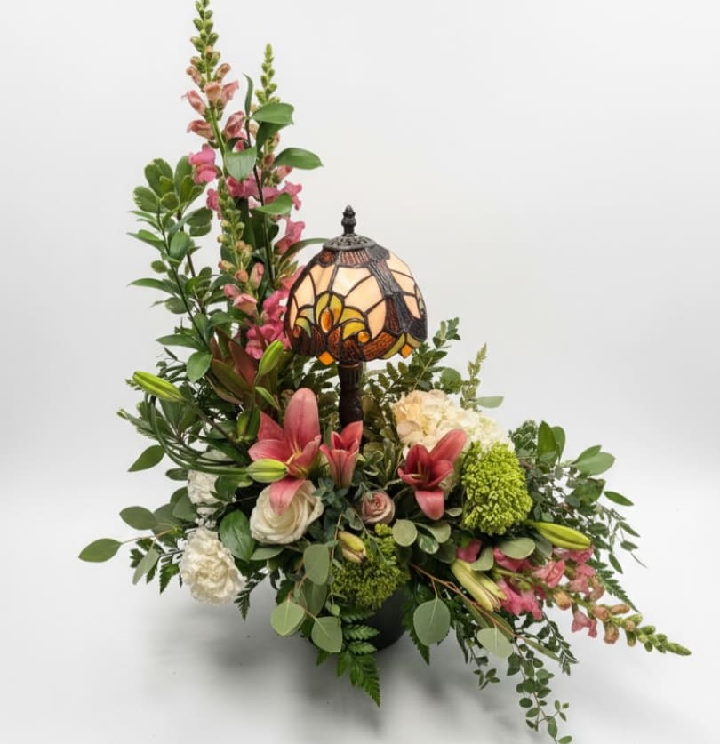 Light of Remembrance - Graceful and comforting, Light of Remembrance is a heartfelt sympathy arrangement designed to honor and celebrate a life beautifully lived. Soft pink lilies, elegant white roses, delicate snapdragons, and lush green hydrangea are thoughtfully arranged with fresh seasonal greenery to create a design that feels both peaceful and uplifting.  At the center of the arrangement sits a beautiful Tiffany-style keepsake lamp, symbolizing warmth, love, and the enduring light of cherished memories. After the flowers have faded, the lamp remains as a meaningful reminder of comfort and remembrance.  Light of Remembrance is a thoughtful gesture for funerals, memorial services, or sending comfort to loved ones during a difficult time.