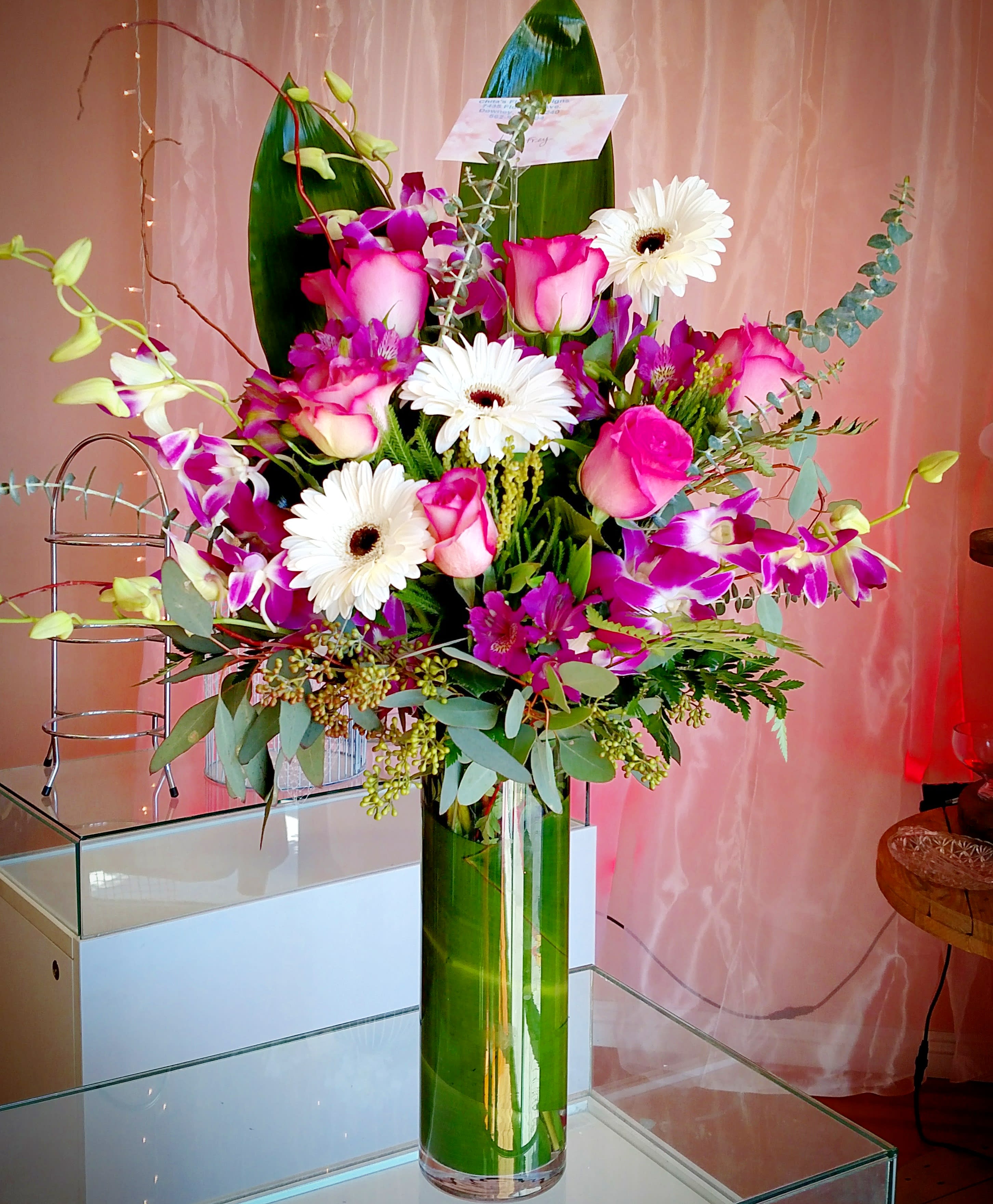  Exotic Bombay Orchids - Tall vase either square or round, with T leaf wrapped inside, filled with Bombay Orchids, roses and Gerber daisies in a one sided design with The leaf backing and curly willow.