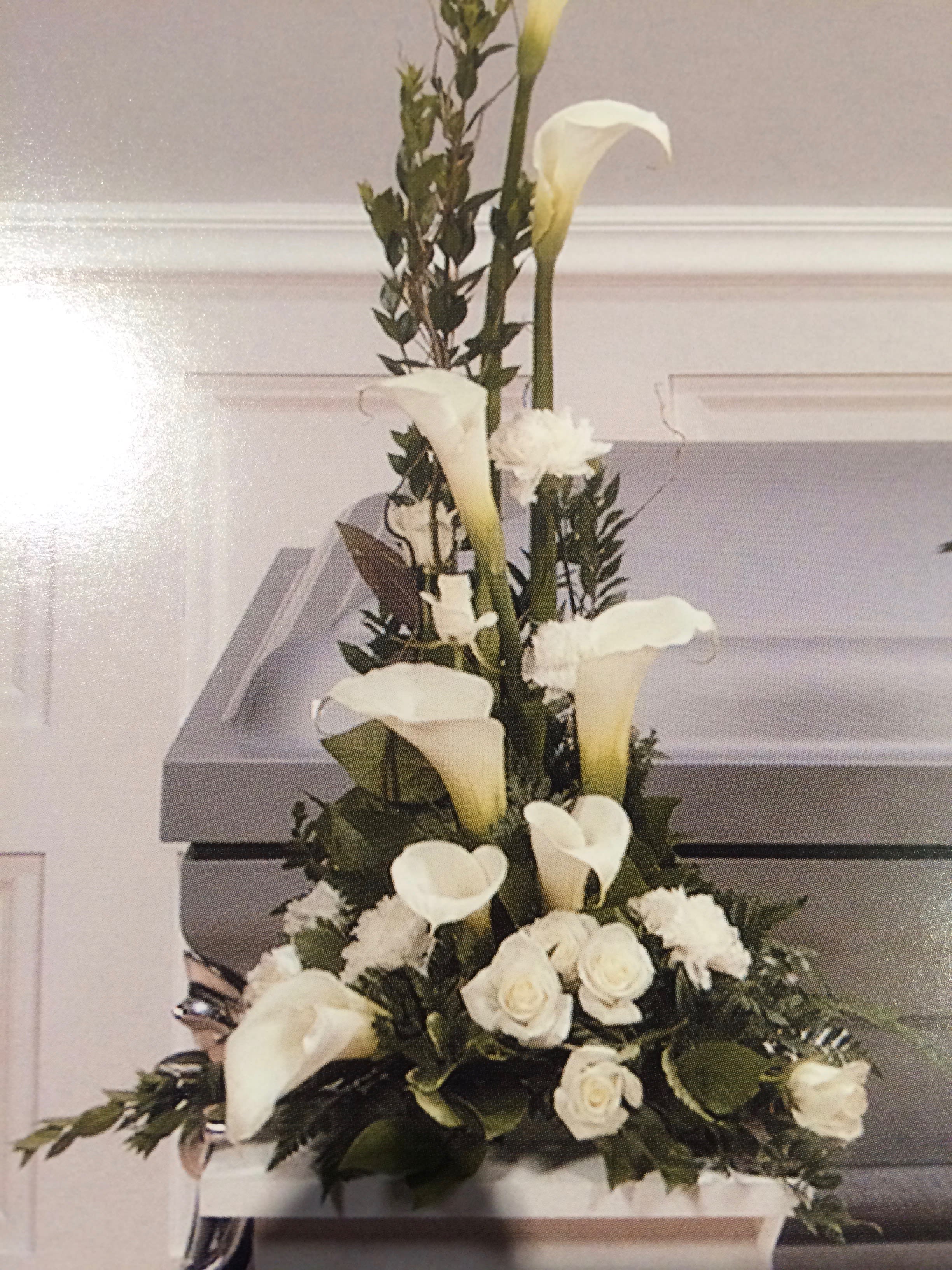 Calla Lilies Pedestal Arrangement  - Show your feelings thru flowers with this Calla Lilies Pedestal arrangement. Alluring calla lilies are paired with a few white carnations &amp; pure white roses with assorted foliage in a designer tray to complete this design. (If you would like a ribbon &amp; wording, i.e., loving friend, Dear Mother, In-Law, Nephew, etc. Please call us for pricing.)