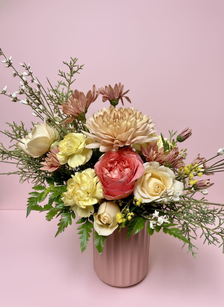 Peach Blossom - This enchanting Valentine's Day arrangement is a beautiful blend of soft pastels and lively blooms, thoughtfully arranged in a light pink ribbed ceramic vase. A stunning pink garden rose is surrounded by the cheerful mimosa, vibrant salmon mums, and delicate white ginestra. Buttery yellow carnations and creamy spray roses add warmth, while the carolina sapphire brings a touch of fresh greenery. The mix of colors and textures creates a romantic and inviting display, making it the perfect way to express love and affection this Valentine's Day.   All flowers, colors, and containers are subject to change due to availability. 