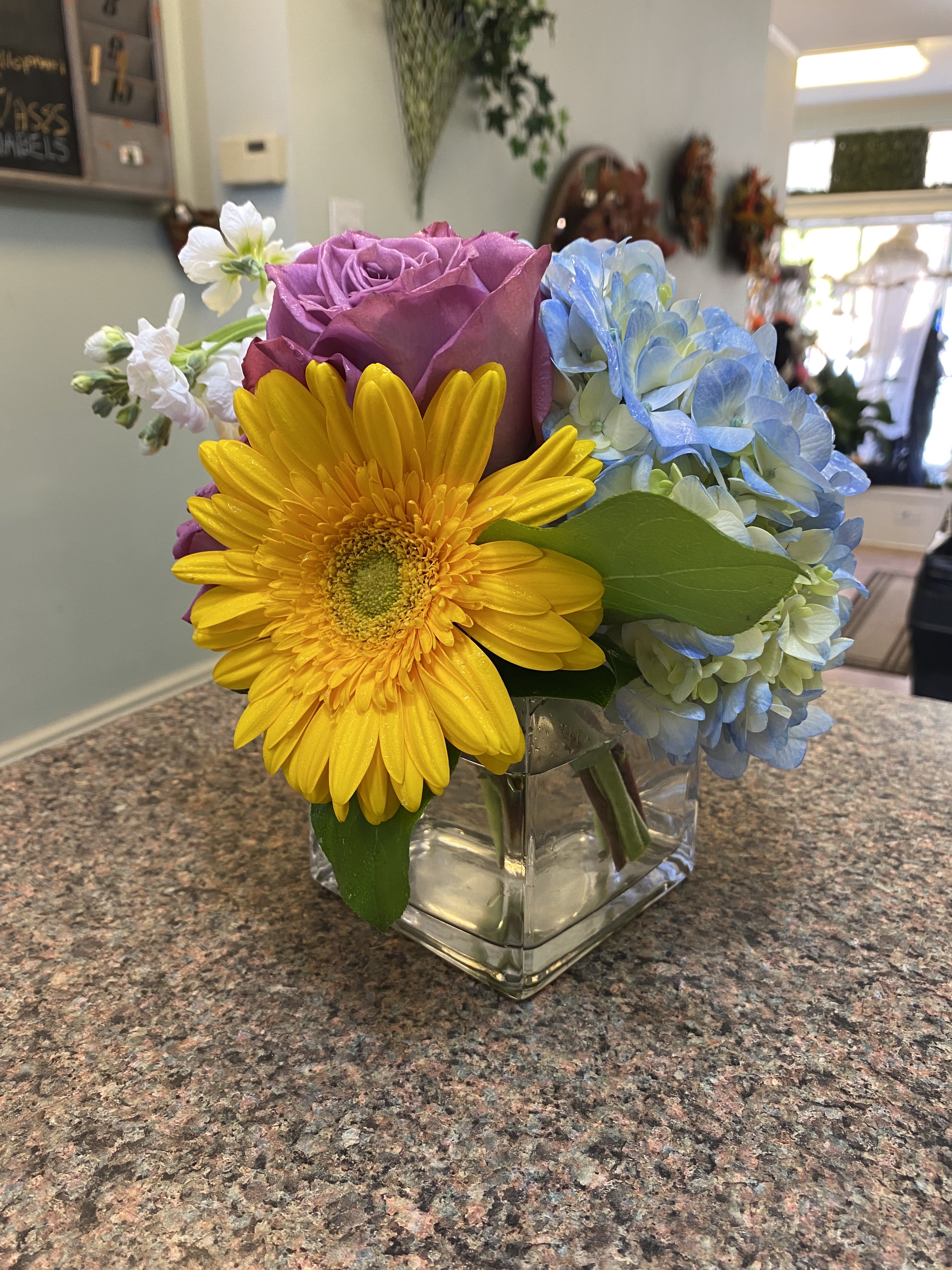 Colorful bouquet with a yellow gerbera daisy, purple rose, and blue hydrangea in a glass vase