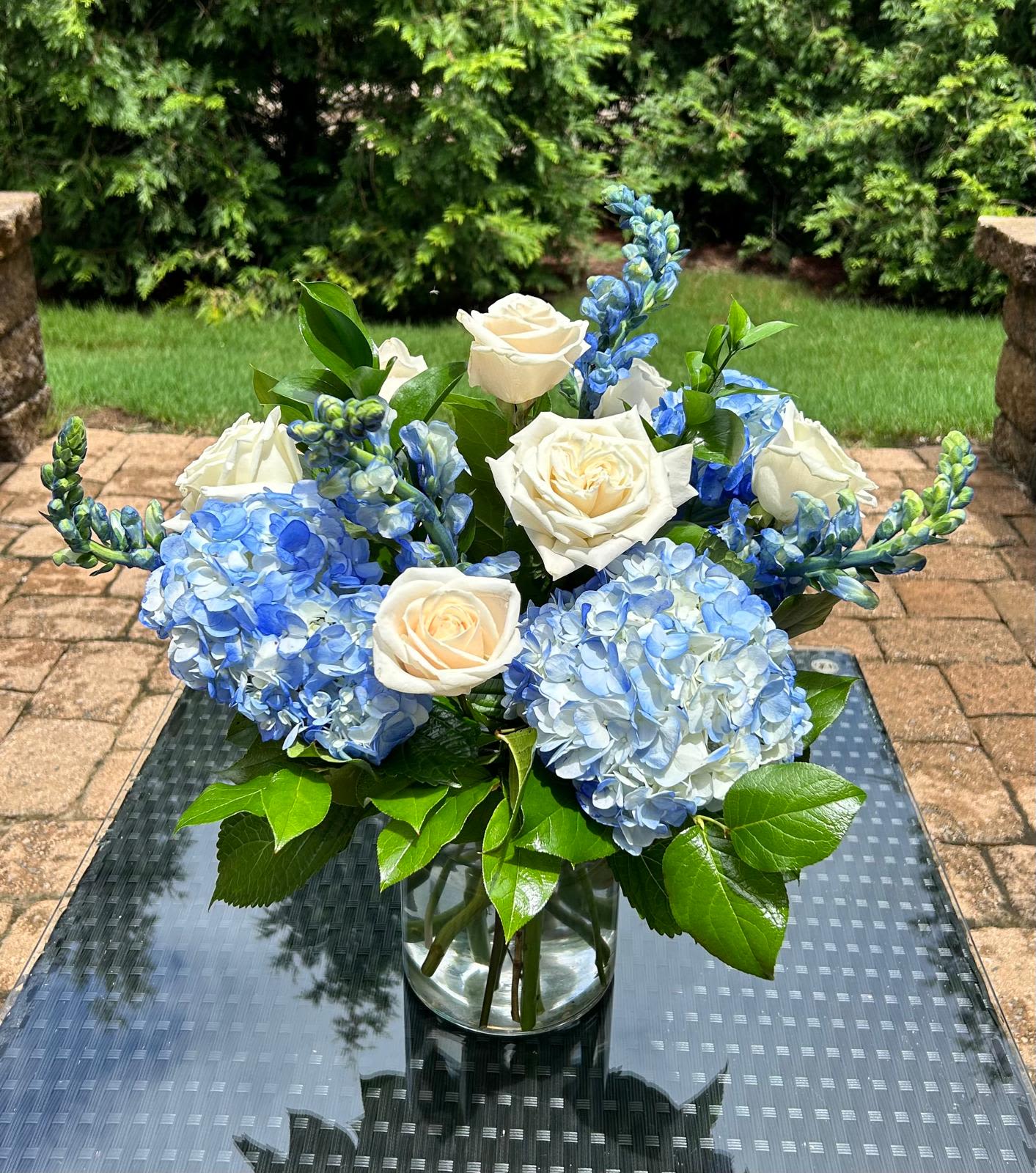 Blue Serenity - Luxury arrangements with real and fresh roses, make this arrangement a true symbol of luxury for all occasions.  Bloom Details: Blue Delphinium, Blue Hydrangea, White Rose Presented by: Crystal Clear Glass Vessel All Occasion  Place your Order Online Monday to Saturday before 1:00 p.m. (E.T.) for same-day delivery.  “Orders received after hours will be delivered the next business day”. Check our coverage area.  Occasionally, flowers and/or container substitutions happen due to weather, seasonality, and market conditions, which may affect availability. If this is the case with the gift you’ve selected, we will ensure that your arrangement's style, theme, and color scheme are preserved and will only substitute items of equal value or higher value.  CARE INSTRUCTIONS	 Keep the flower arrangement hydrated with natural water and change the water every two days. Do not spray any cleaning product on the flower arrangement. Do not place heavy objects directly on the flower arrangement. Keep flower arrangements out of direct sunlight and extreme heat. 
