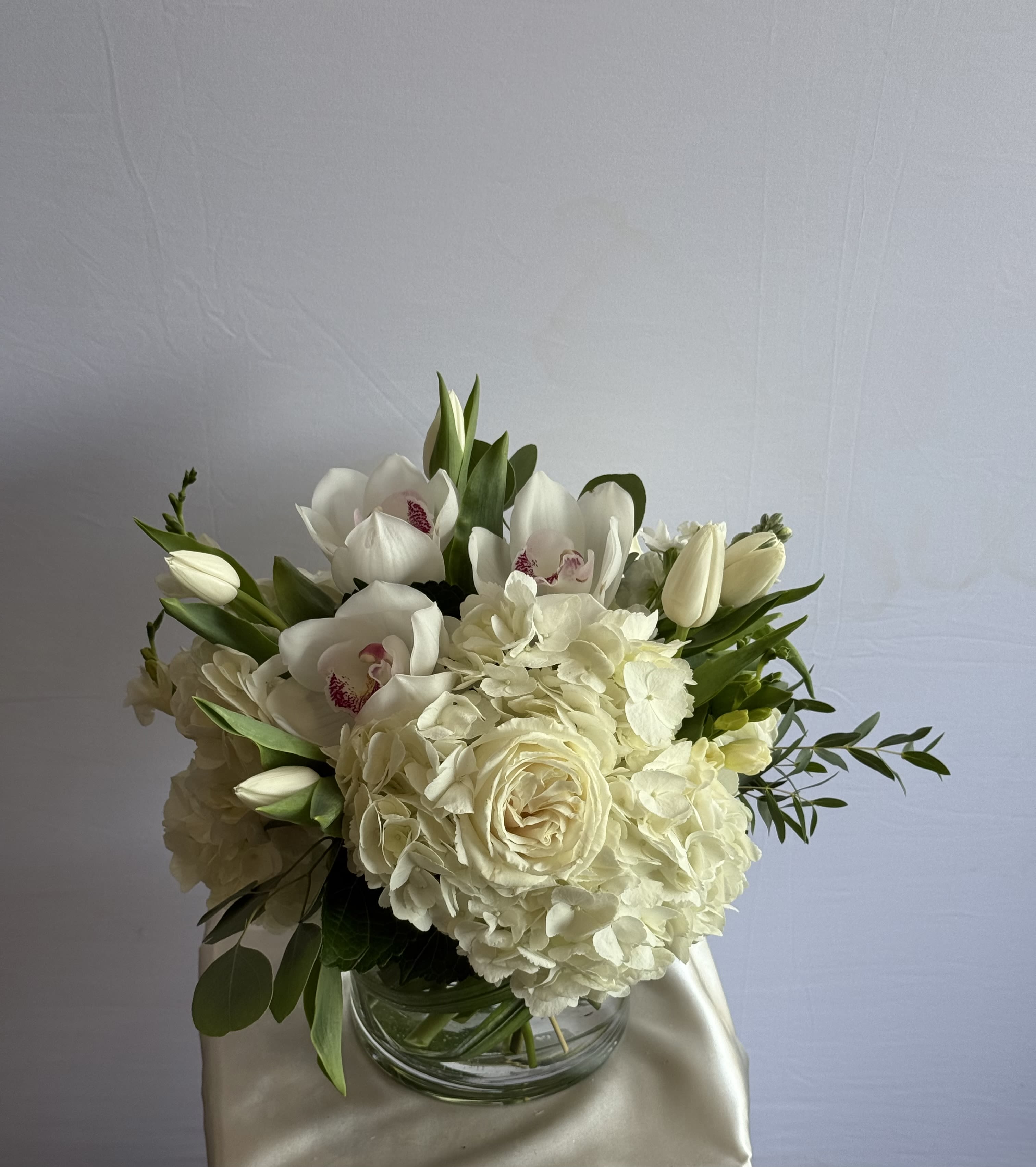Snowfall Serenity - Graceful, timeless, and radiating refined charm, snowfall serenity is a monochromatic masterpiece that brings together the purest shades of white in a luxurious floral symphony. This compact yet lush arrangement features opulent white hydrangeas, velvety garden roses, and exotic white cymbidium orchids, accented with elegant white tulips and lush greenery. 