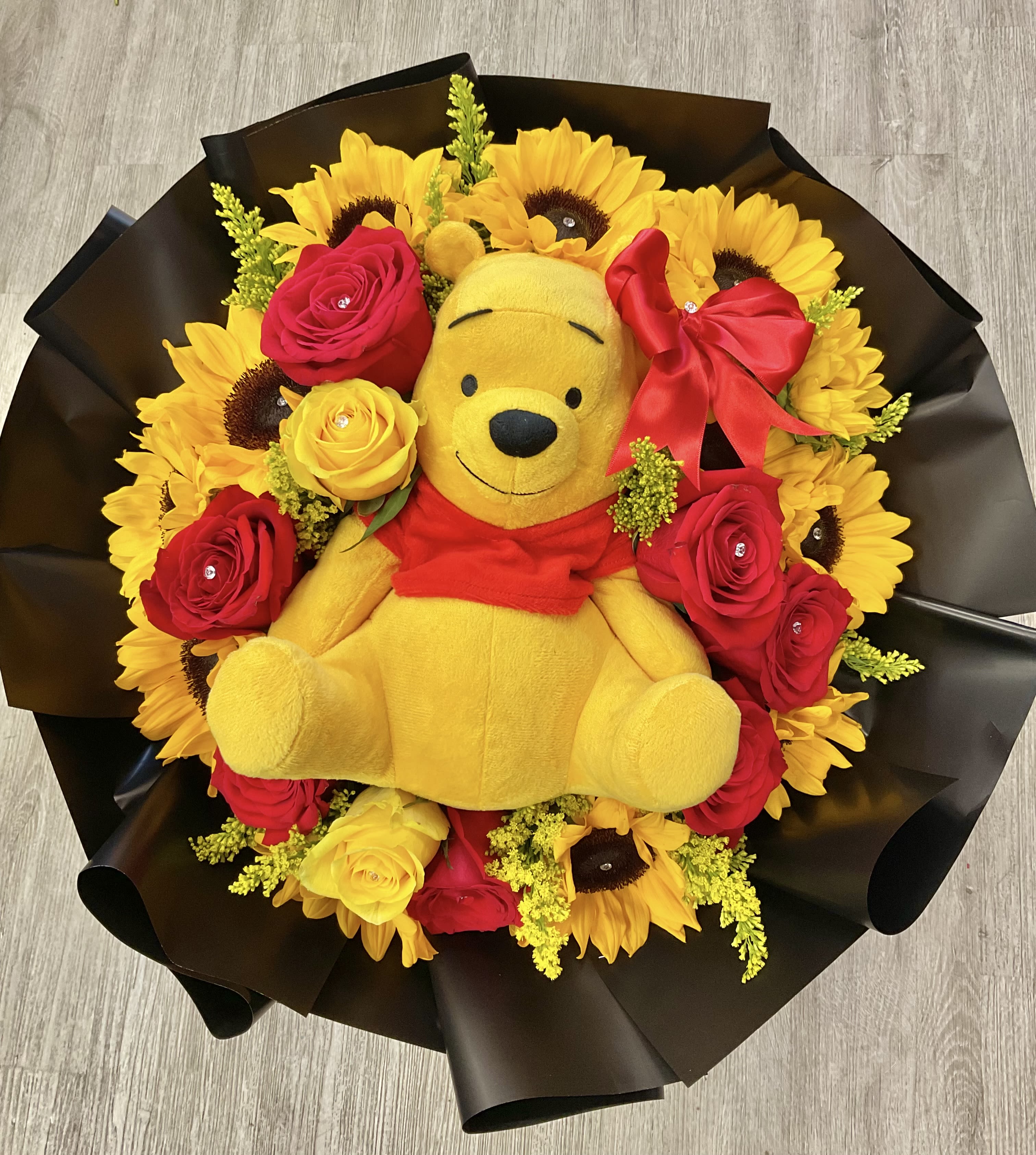Pooh's Garden Of Joy - This charming floral bouquet that combines the bright and cheerful beauty of sunflowers, and the romantic and elegance of roses, creating a perfect balance of warmth and love. The combination of these flowers, paired with this beloved bear, makes this bouquet an enchanting gift, for any occasion that calls for a little extra sweetness and cheer. 