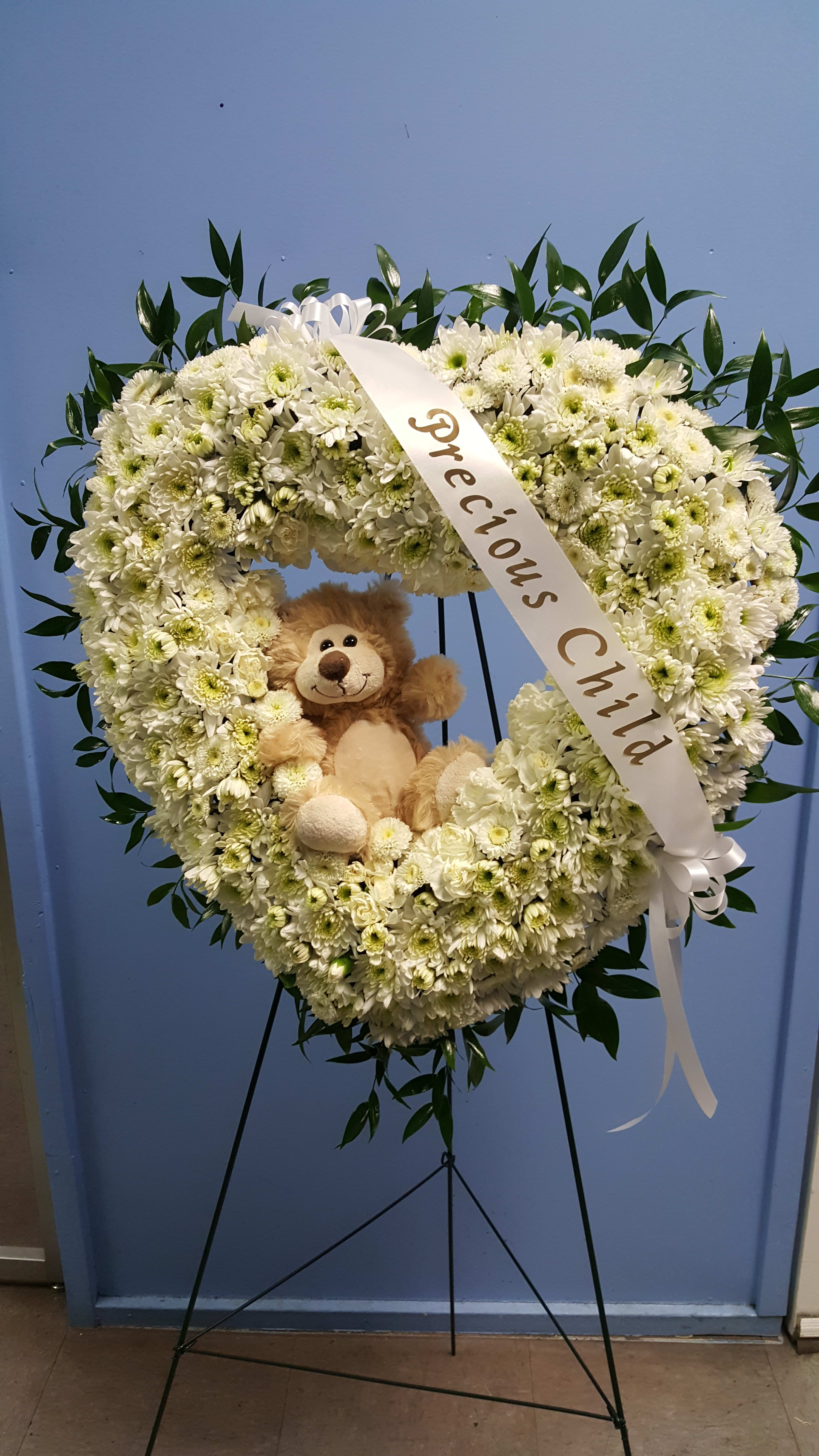 Heart-shaped white flower wreath with a teddy bear and ribbon on a stand