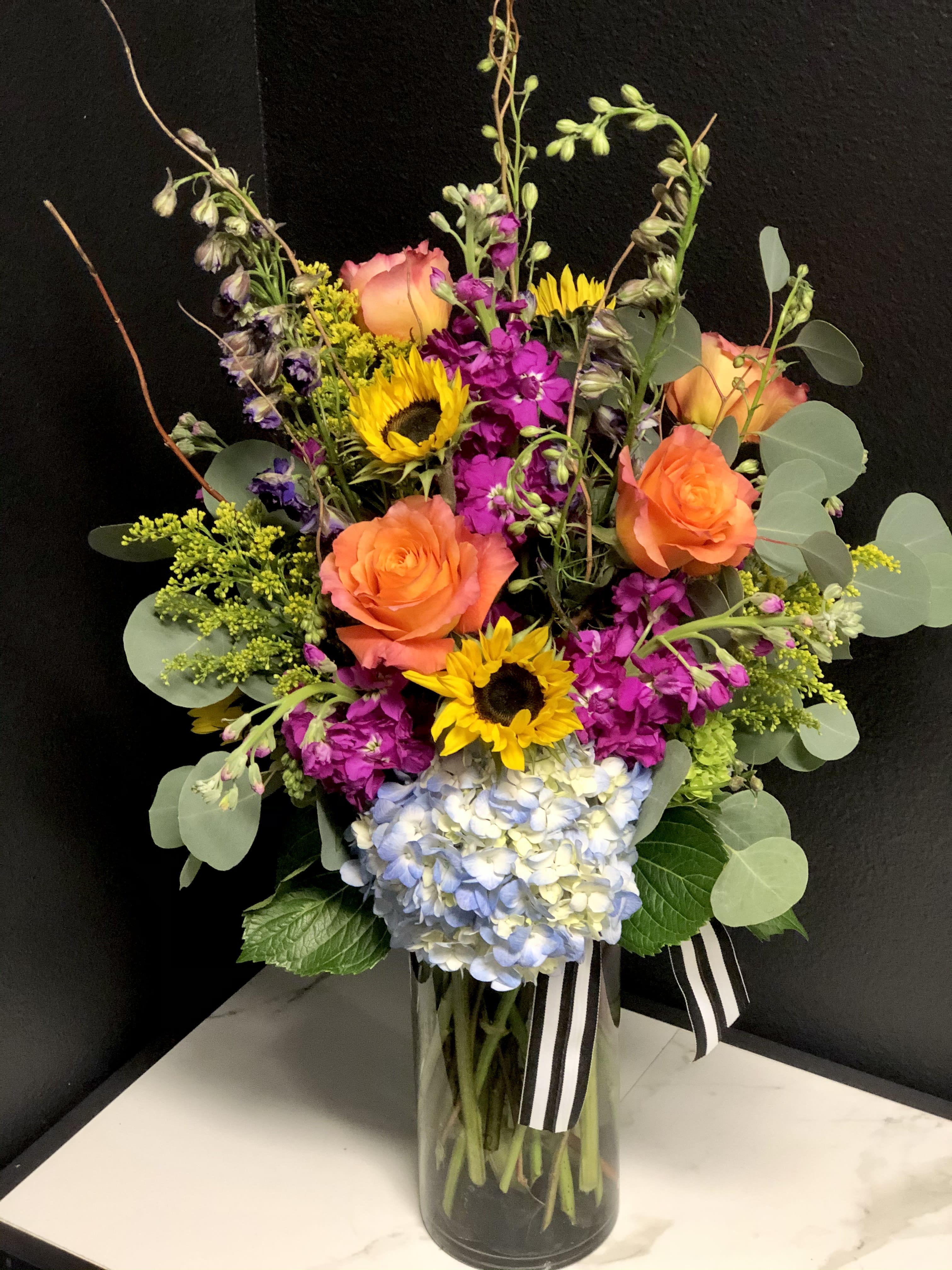 Tall Vibrant Garden  - A bright elegant garden of Sunflowers, Hydrangea, Stock, Roses, and seasonal foliage. Designed in a clear glass vase. Approximately 24"H x 15"W  Design will increase in overall size and stem count in the Deluxe &amp; Premium Options. 