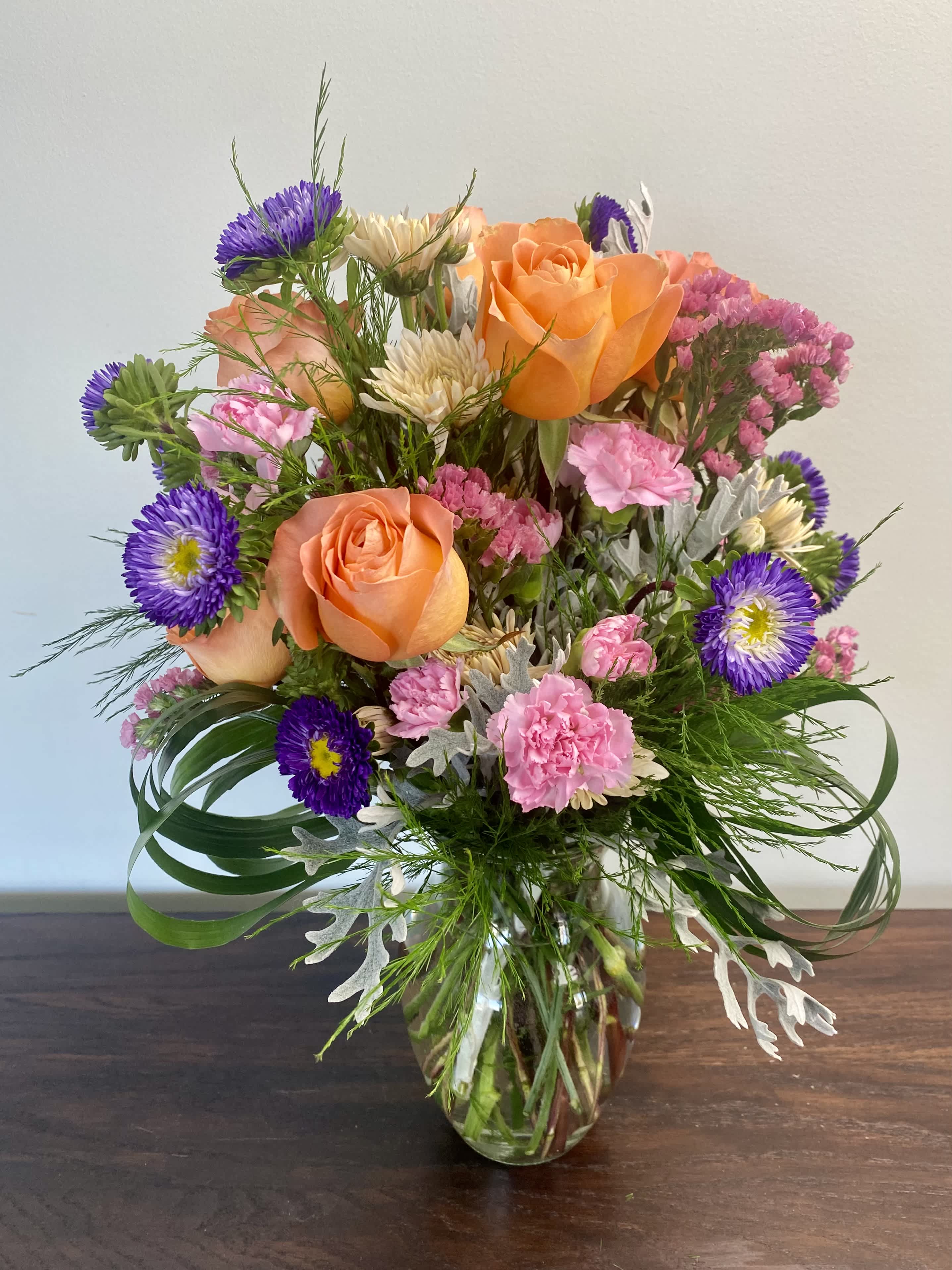 Joyful Blossoms - A bright and cheery design sure to bring a smile to the receiver! Including Sunset Roses, Purple Matusomtos, Champaign Cremons, Pink Carnations, and mixed greens in a glass vase. 
