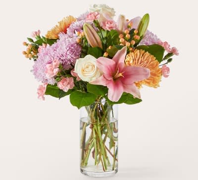 Gelato Bouquet - Bright, joyful, and full of personality, this bouquet is a celebration of color and texture. Soft white roses and peach carnations bring gentle elegance, while vibrant lilies, bold chrysanthemums, and cheerful marigold tones add a lively pop that instantly lifts the mood. Accents of orange berries and lush greenery tie everything together, creating a rich, garden‑fresh look that feels both modern and heartfelt. Arranged in a clear glass vase, it’s the perfect way to send warmth, gratitude, or a burst of happiness to someone special.