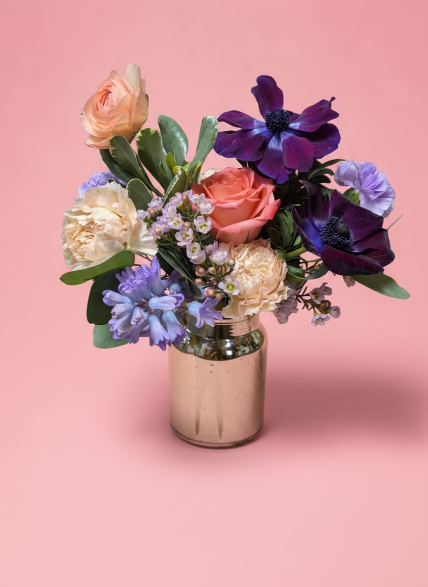 Champagne Taste - Celebrate love in style with Champagne Taste, an arrangement that’s equal parts elegance and whimsy. Nestled in a shimmering champagne mercury vase, this bouquet features regal purple anemone, vibrant coral ranunculus, soft lavender moonshade carnations, playful silly putty carnations, fragrant lavender hyacinth, a romantic coral rose, and delicate pink waxflower.  Perfect for someone with a taste for the finer things, this arrangement combines luxe sparkle, bold color, and soft romance—making it a truly unforgettable gift.   All flowers, floral colors, and containers are subject to change due to availability.