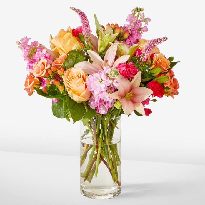 The Bisous Bouquet - Bursting with color and personality, this bouquet is designed to brighten any moment. Peach and pink roses bring soft elegance, while vibrant lilies, stock, and mini carnations add layers of texture and cheerful energy. Accents of hypericum berries and lush greenery create a full, garden‑fresh look that feels both uplifting and refined. Arranged in a clear glass vase, it’s a radiant display that instantly warms a room.