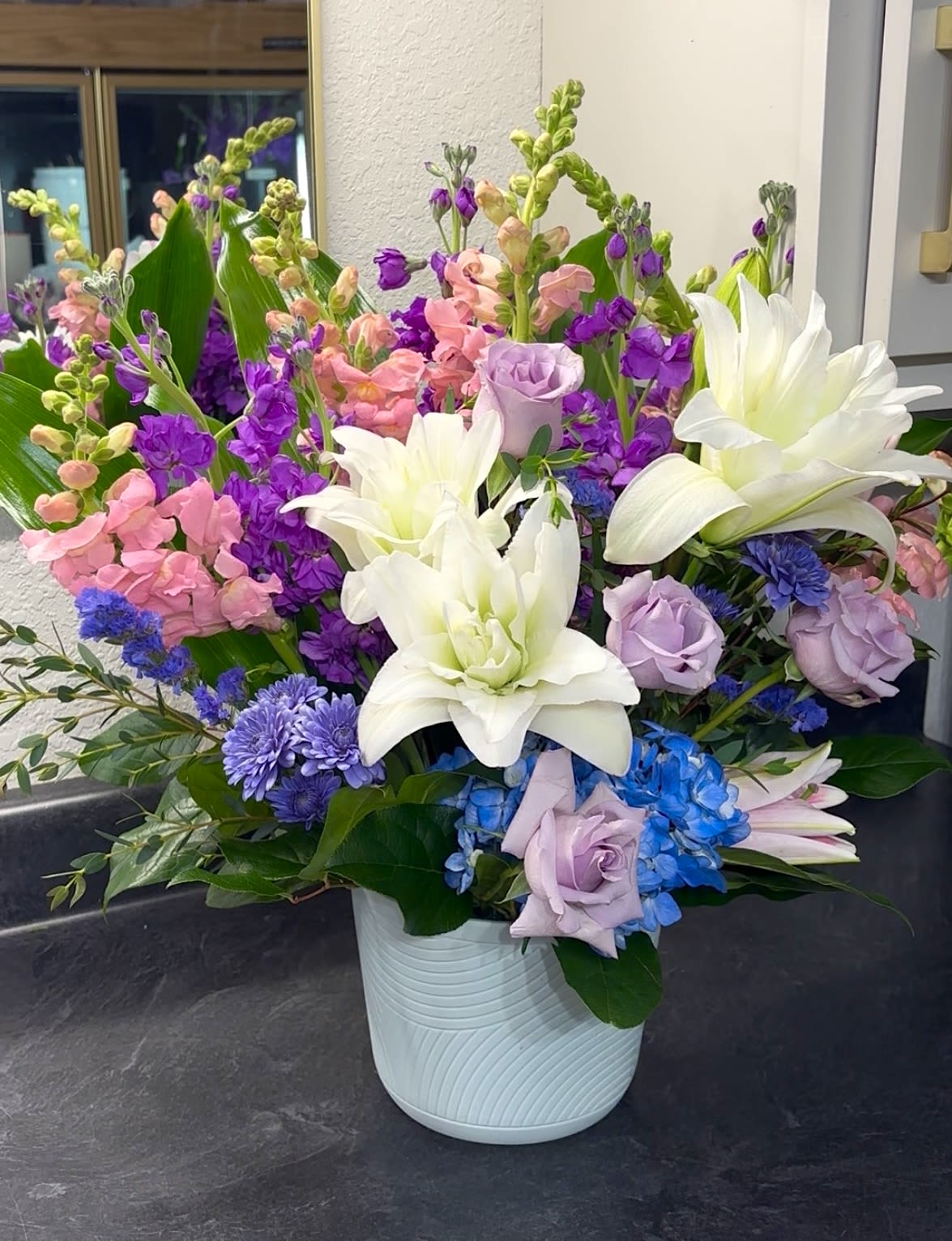 Blooming Elegance - Bright and colorful blooms perfect for any occasion. Includes white Double Lilies, purple Roses, purple Stock, pink Snap Dragons, blue Hydrangea, and purple Daisy Poms in a ceramic vase. 