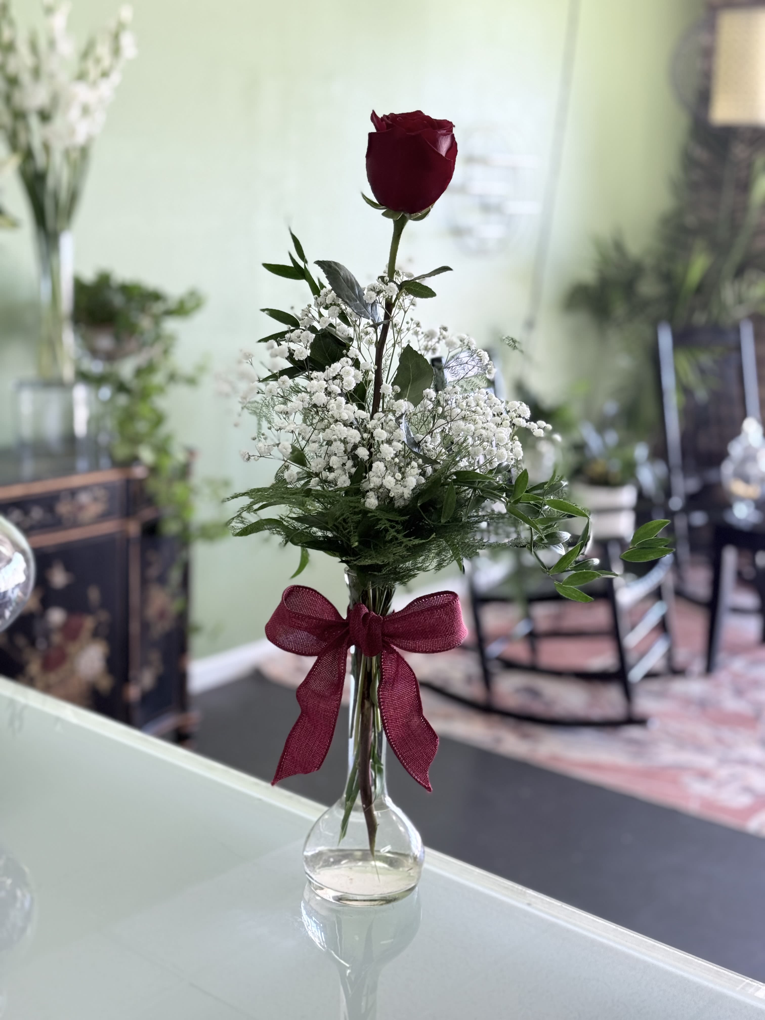 Single red rose in a clear glass vase with white baby's breath and a burgundy ribbon