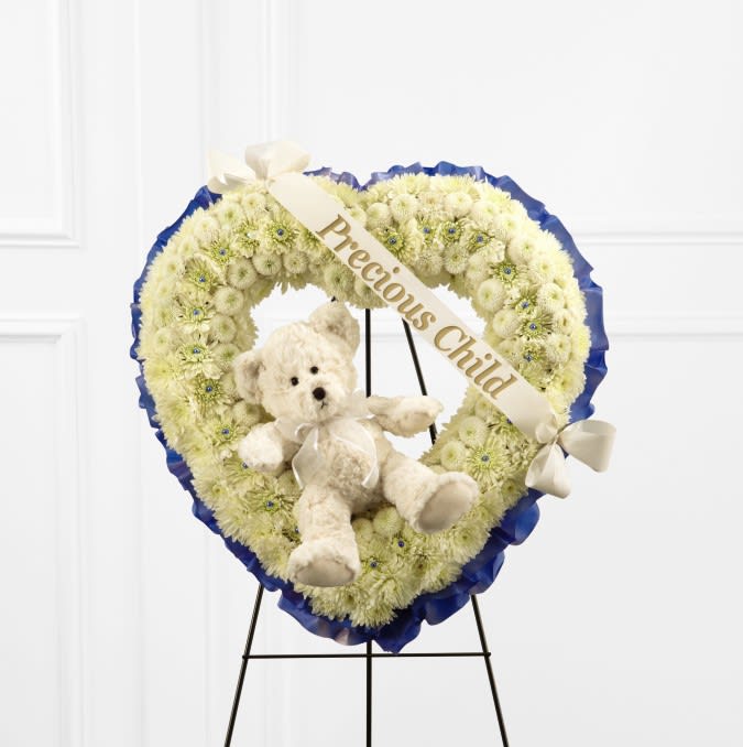 The FTD® Precious Child™ Standing Heart  - Beautiful and pure white flowers commemorate a life lost too soon. White pompons are designed by a local florist in a touching heart shape to create our Precious Child™ Standing Heart. Each white bloom is accented by a satin blue ribbon outline, an ivory plush bear and a printed ribbon.  Details: o Heart spray is approximately 22"H x 20"W 